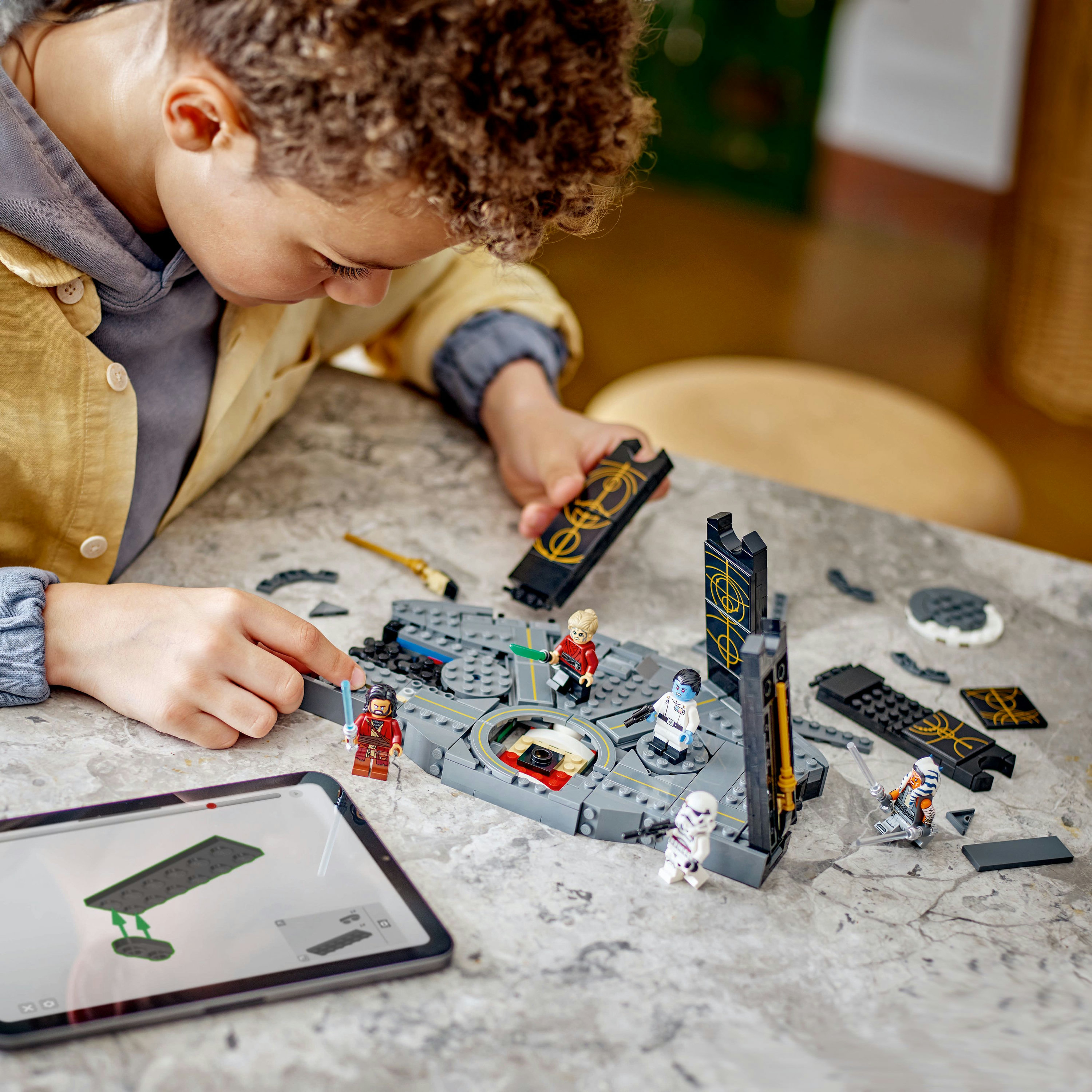 Un bambino costruisce un set Lego Star Wars. La scena comprende personaggi Lego, un tablet e vari pezzi Lego su una superficie grigia.