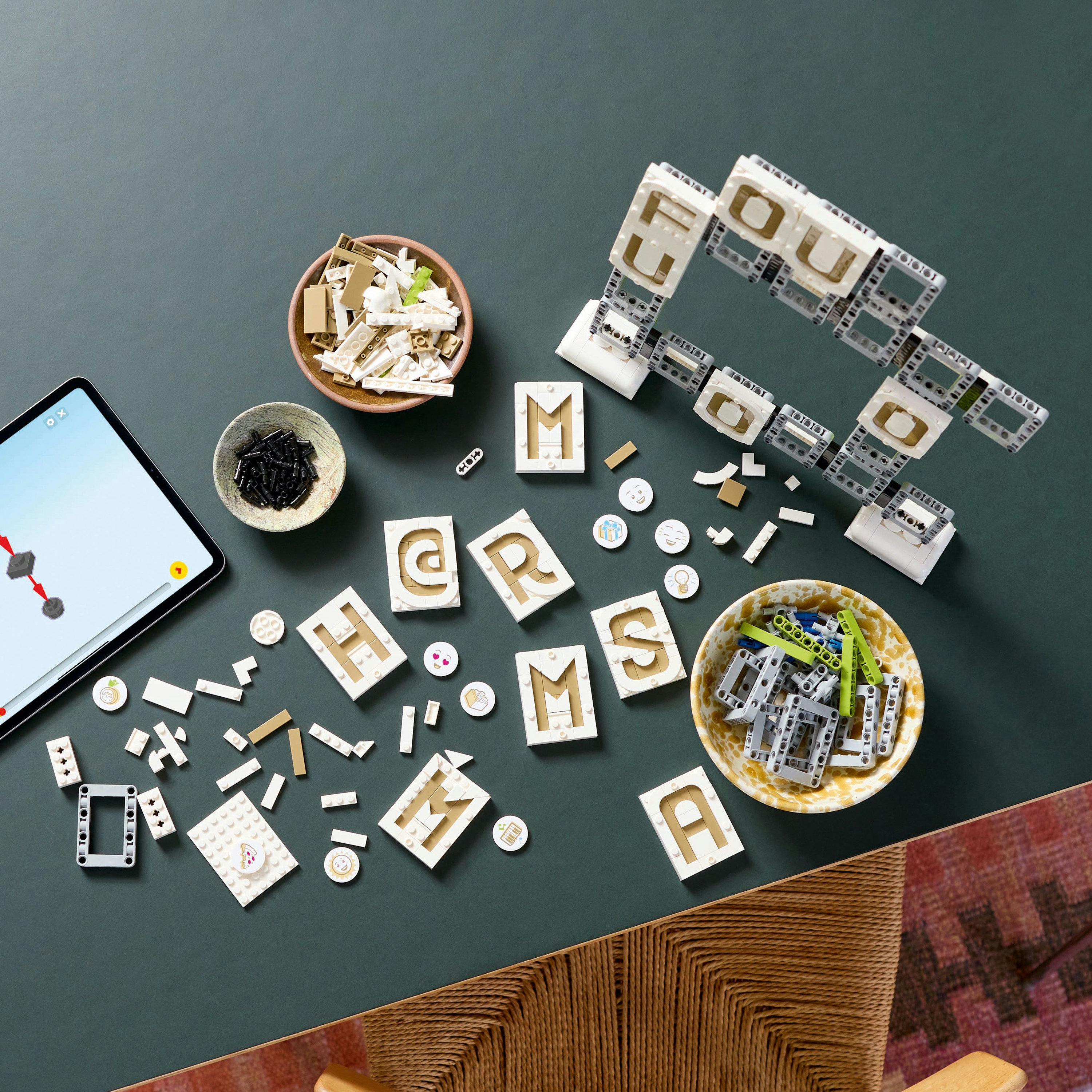 LEGO tuğlaları, kaseler ve bir tablet bulunan bir masa. LEGO'lar "HERMS" ve "YOU" kelimelerini heceliyor. Çeşitli şekiller ve renkler mevcut.
