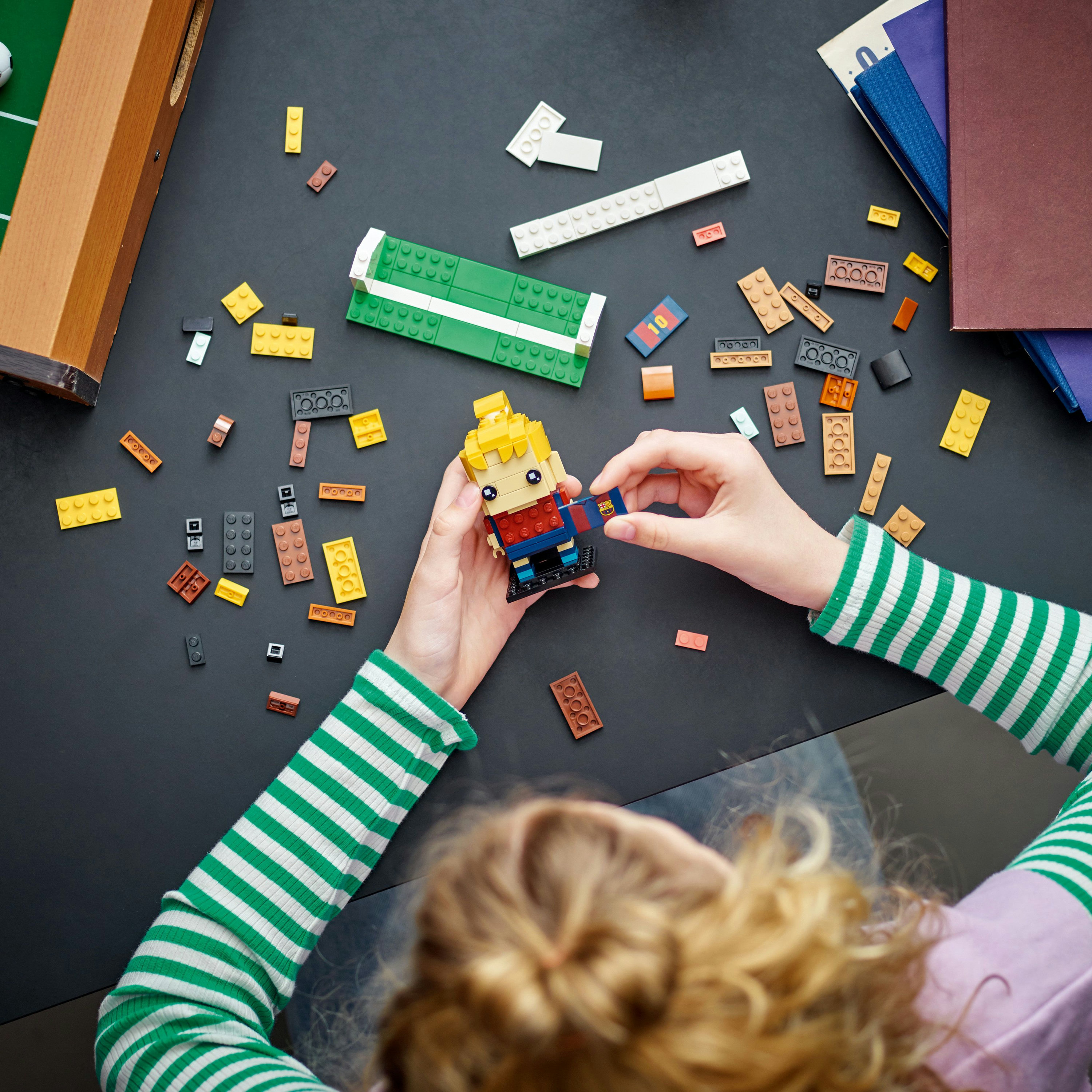 Bir kişi siyah bir masa üzerinde bir Lego figürü inşa ediyor. Yeşil ve beyaz çizgili kol görünüyor. Diğer Lego parçaları dağınık.