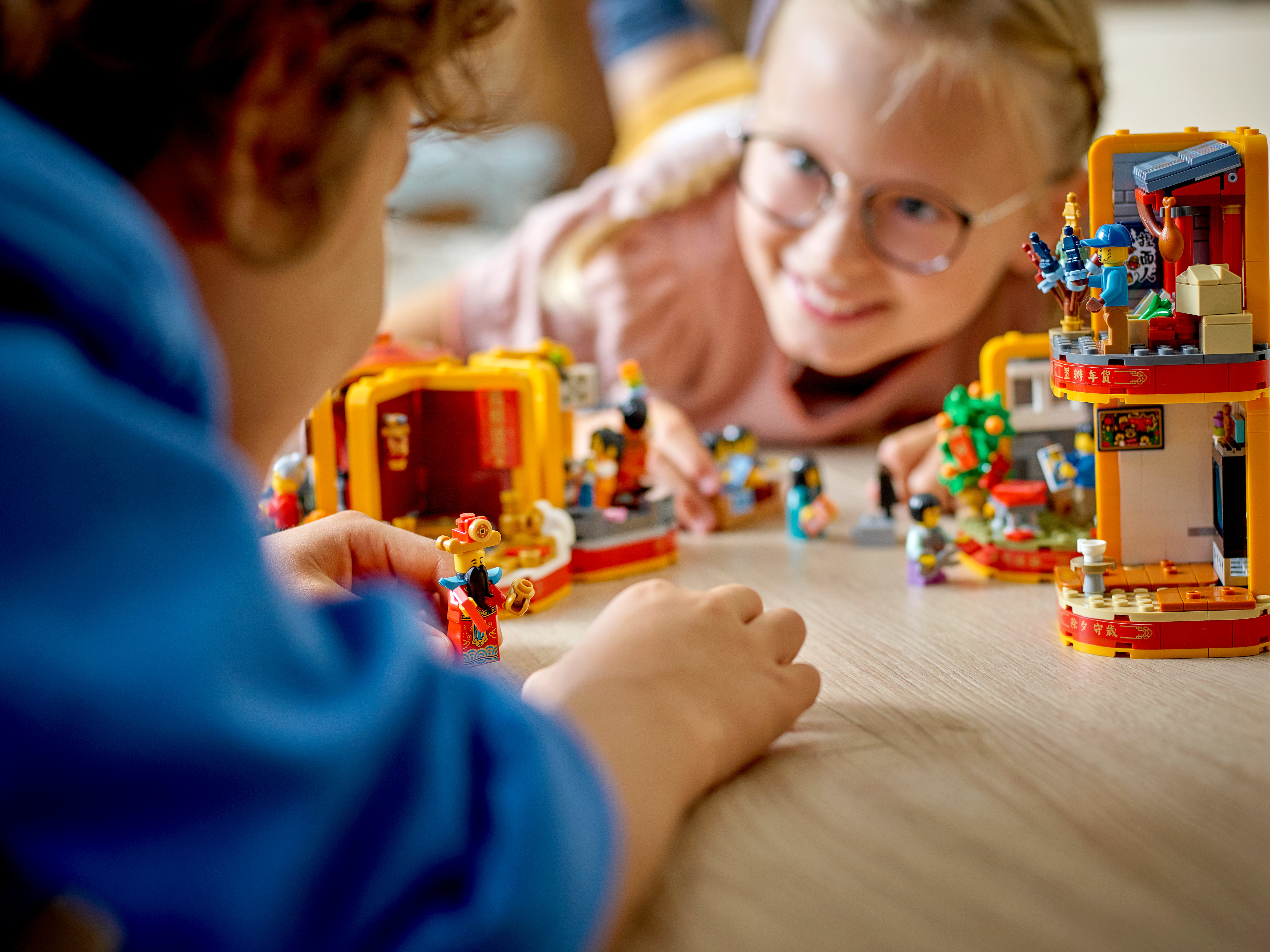 Dwoje dzieci bawiących się zestawami Lego na drewnianej powierzchni. Jedno jest na pierwszym planie. Drugie uśmiecha się w tle.