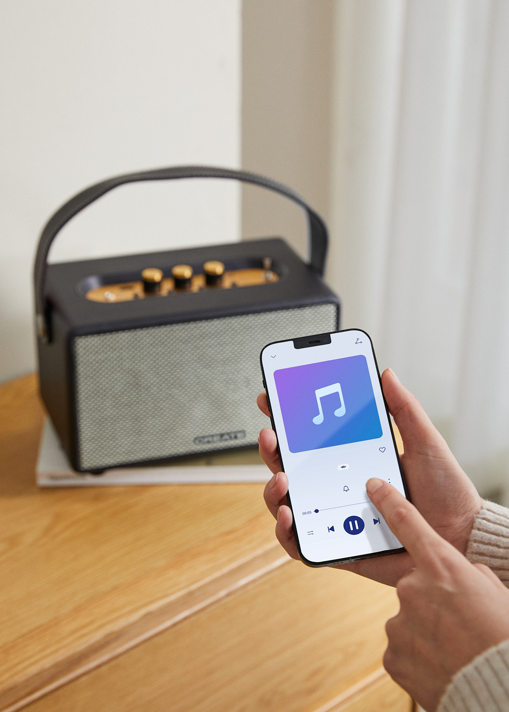 Una persona sostiene un teléfono inteligente que muestra una aplicación de música. Un altavoz está sobre una mesa, junto a una pila de libros. La habitación tiene una pared blanca y una cortina.