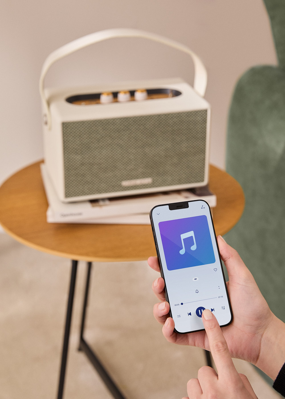 Una mano sostiene un teléfono reproduciendo música, un altavoz está sobre una mesa, y un sillón en el fondo.