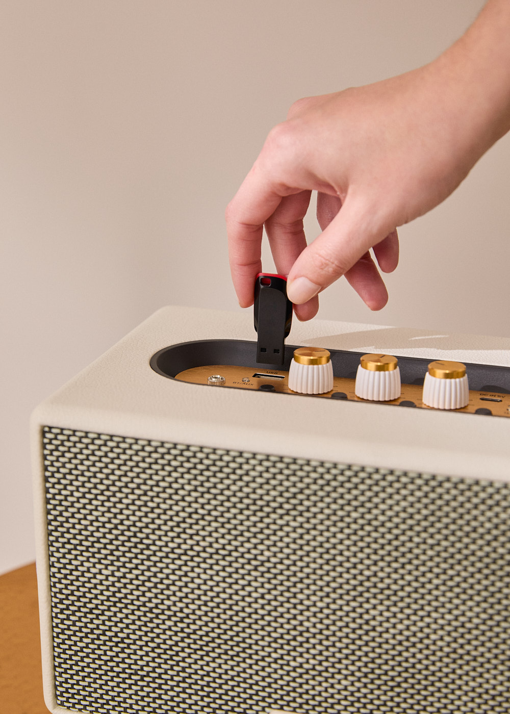 Una mano inserta una unidad USB en un altavoz. El altavoz es blanquecino con detalles dorados. El fondo es beige liso.