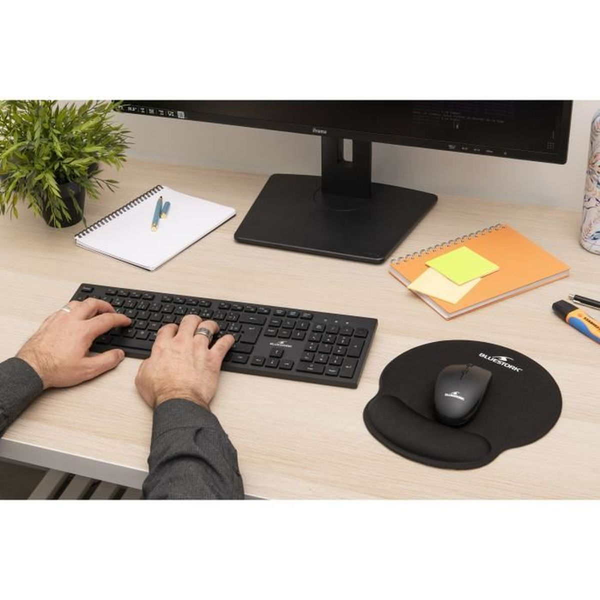 Hände tippen auf einer Tastatur auf einem Holztisch. Monitor, Notizbücher, Maus, Stift und eine kleine Pflanze sind ebenfalls zu sehen.