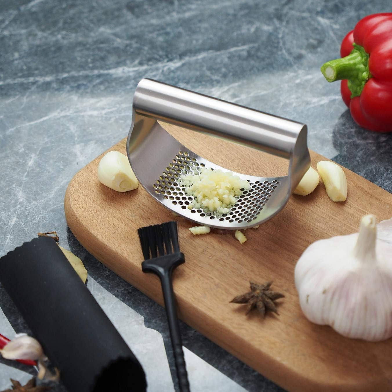Eine Knoblauchpresse mit gepresstem Knoblauch, Knoblauchzehen, einer Knolle und einer roten Paprika auf einem Holzbrett.