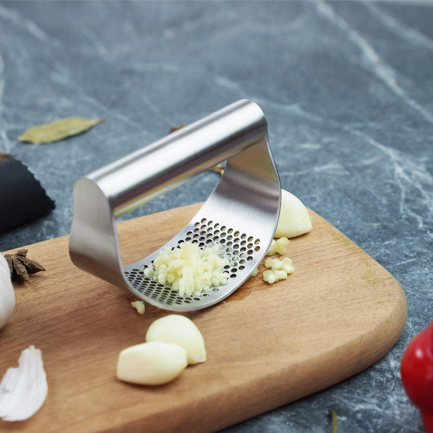Eine Knoblauchpresse mit gepresstem Knoblauch, Knoblauchzehen und einem Holzbrett. Gewürze und ein Marmorhintergrund sind ebenfalls sichtbar.