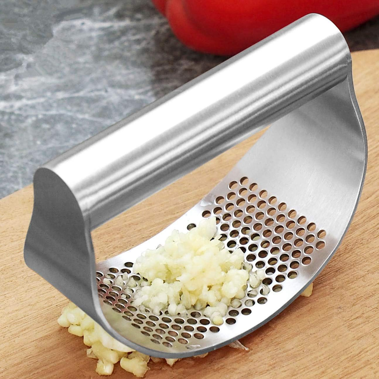 Eine silberne Knoblauchpresse mit gehacktem Knoblauch auf einem Holzbrett, im Hintergrund eine rote Paprika.