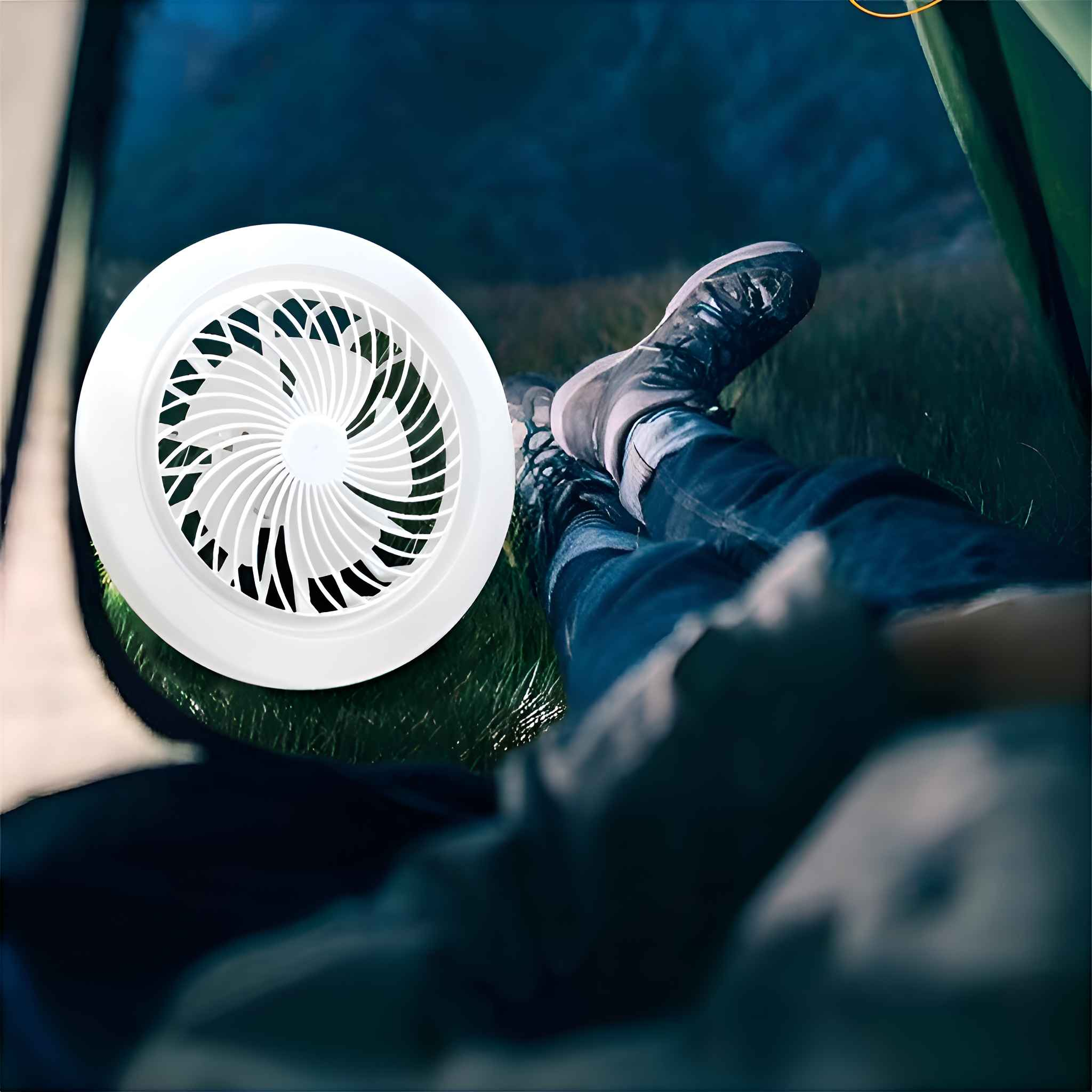 Ein weißer Ventilator in einem Zelt. Beine in Jeans und Schuhen im Vordergrund. Hintergrund: grünes Gras und dunkle, verschwommene Landschaft.