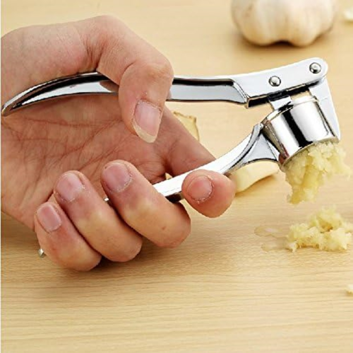 Eine Hand drückt eine Knoblauchpresse und extrudiert gehackten Knoblauch. Eine ganze Knoblauchzehe und -zehen sind im Hintergrund auf einer Holzoberfläche.