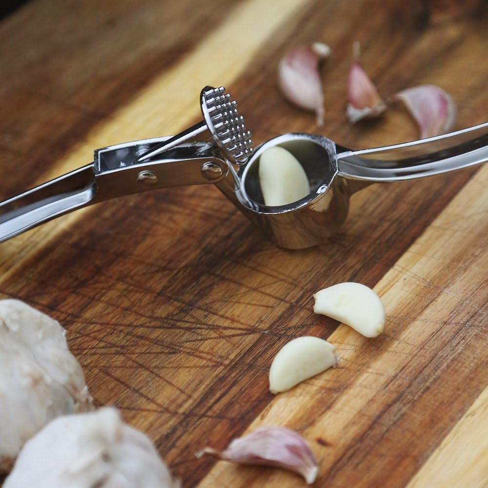 Eine Knoblauchpresse mit einer Knoblauchzehe im Inneren liegt auf einem Holzschneidebrett. Knoblauchzehen sind verstreut.