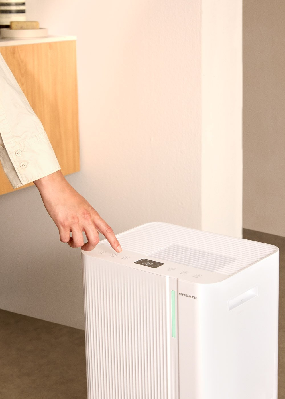 Una mano presiona un botón en un purificador de aire blanco. Un gabinete de madera y una pared blanca están en el fondo.