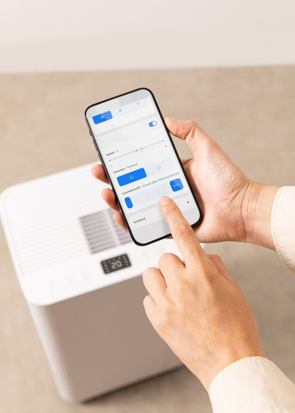 Persona usando un teléfono para controlar un purificador de aire blanco. El teléfono muestra configuraciones de velocidad, dirección y temporizador. La pantalla muestra 20.