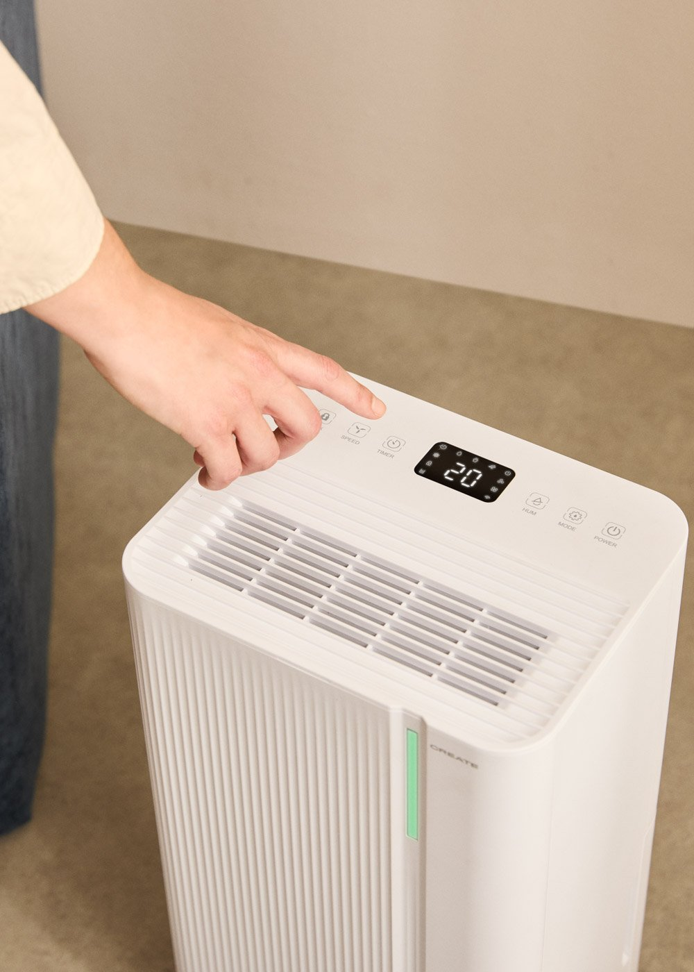 Una mano presiona un botón en un purificador de aire blanco. La pantalla muestra 20. El dispositivo está en una habitación con paredes beiges y suelo gris.