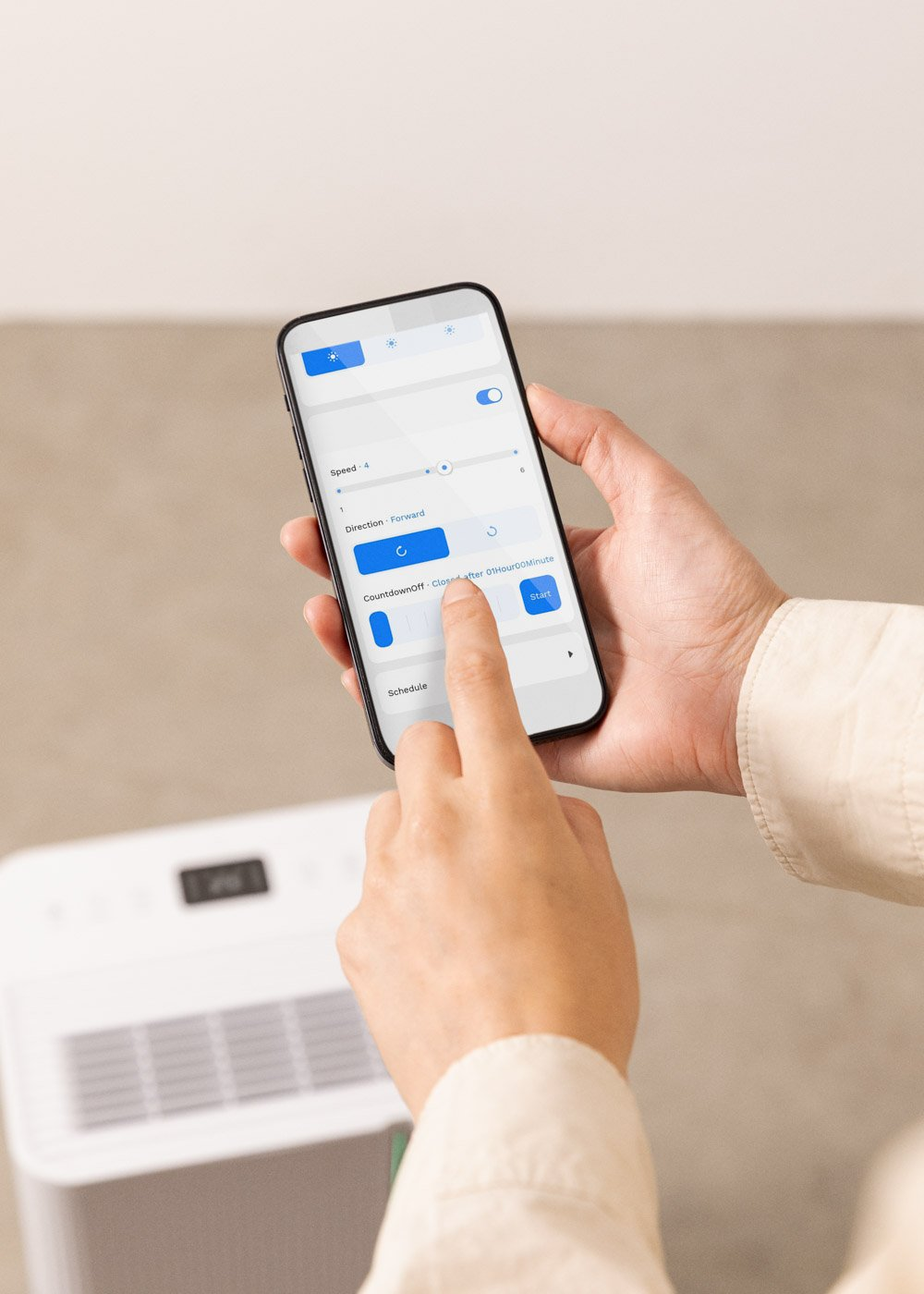 Una persona sostiene un teléfono con una interfaz de aplicación. La pantalla muestra ajustes de velocidad, dirección y un temporizador de cuenta regresiva. Debajo hay un aparato blanco.