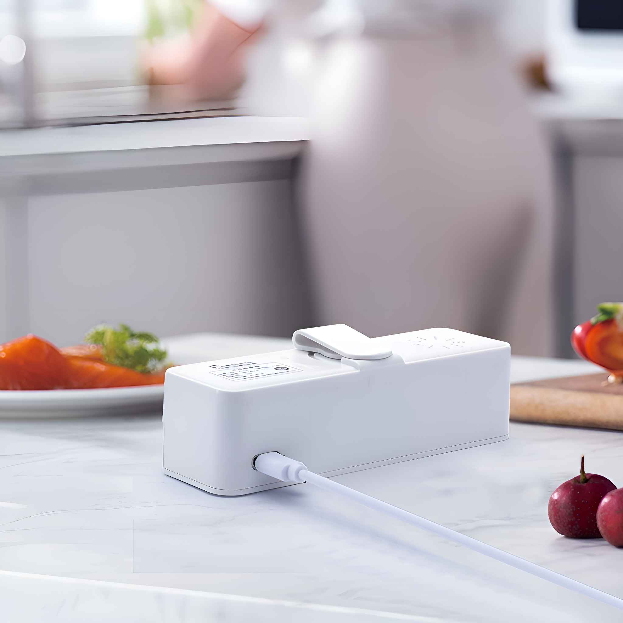 Ein weißes Gerät mit weißem Kabel liegt auf einer Marmoroberfläche. Lachs und Äpfel sind ebenfalls vorhanden. Eine Person im Hintergrund.