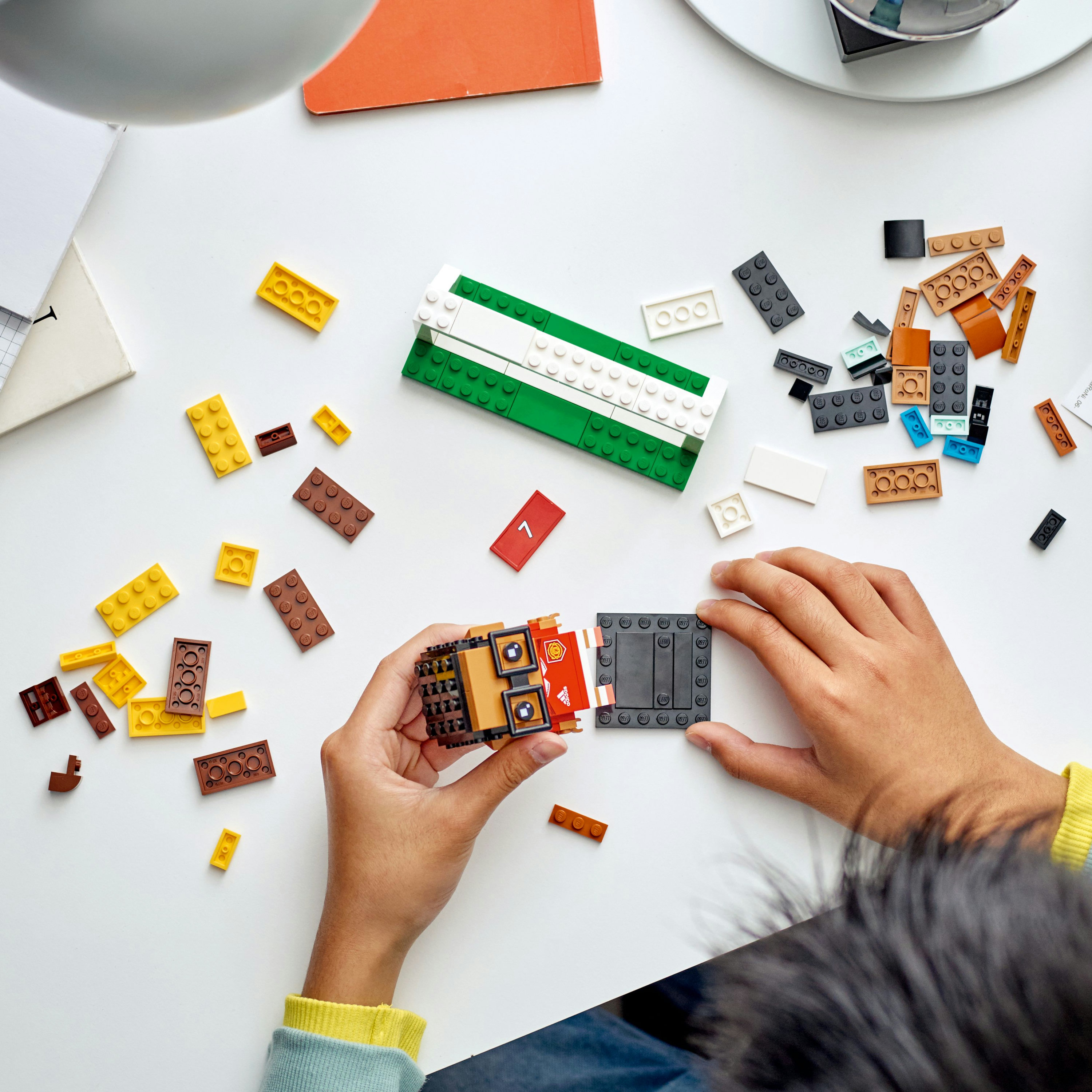 Eller beyaz bir yüzeyde Lego bloklarını birleştiriyor. Çeşitli renklerde bloklar ve kısmen inşa edilmiş bir yapı görülebilir.
