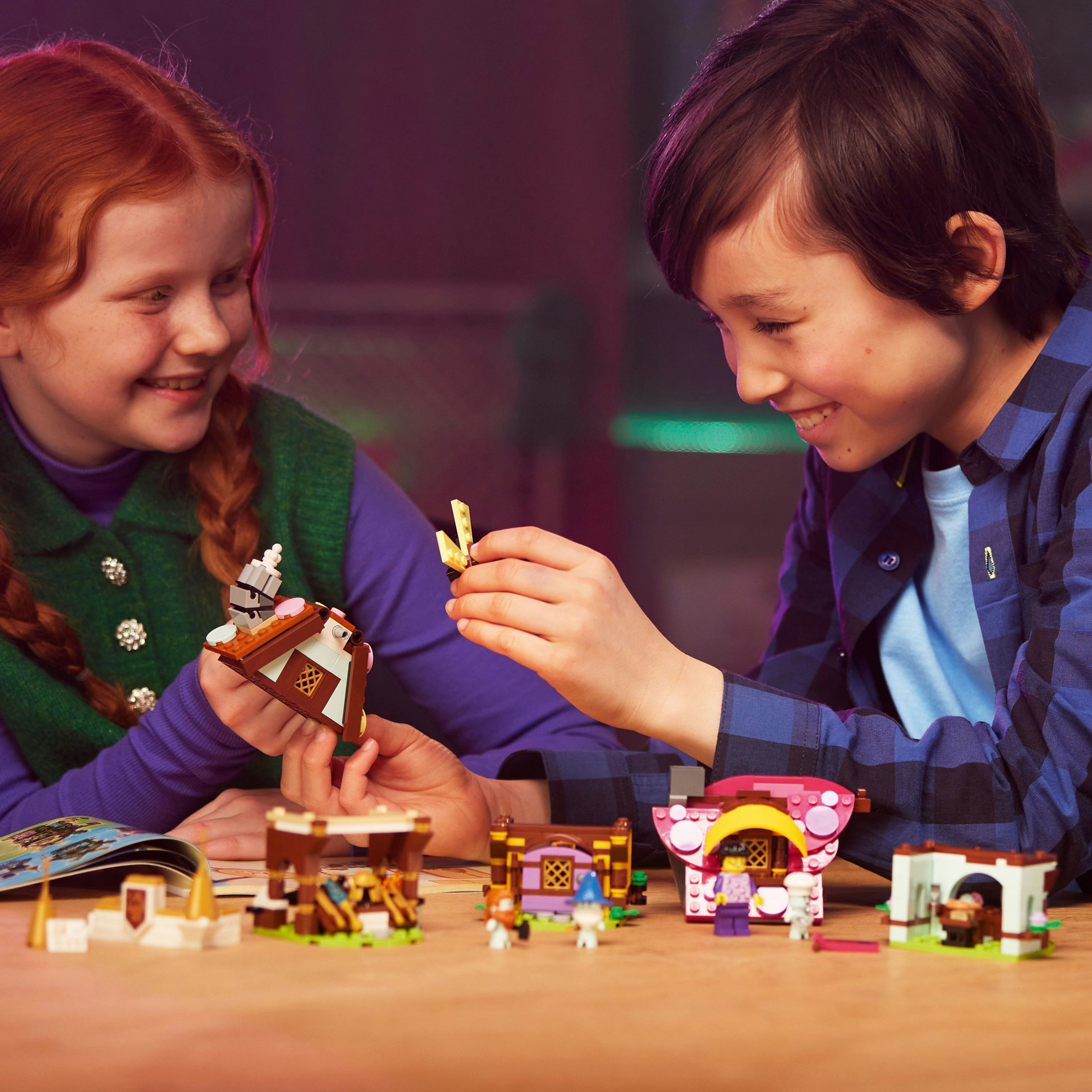 Dwoje dzieci uśmiecha się podczas budowania zestawów Lego. Na stole znajduje się kilka konstrukcji Lego. Jedno dziecko trzyma element Lego.