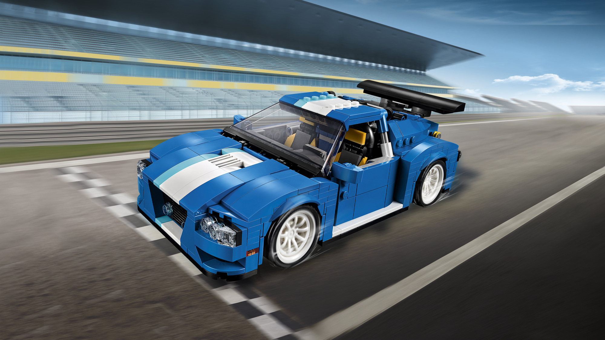 Niebieski samochód wyścigowy Lego jedzie po torze. Białe i niebieskie paski na masce. Trybuny i niebo w tle.