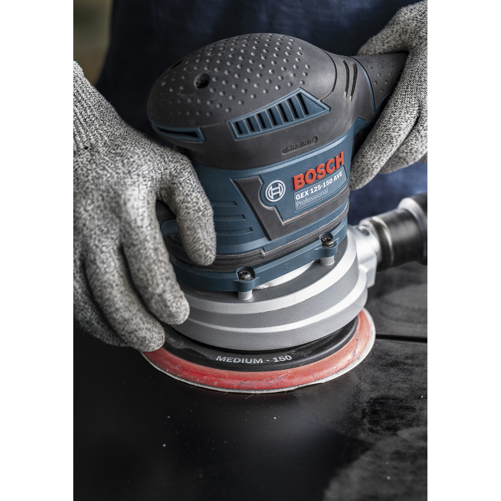 Hände in Arbeitshandschuhen halten einen Bosch GEX 125-150 AVE Schleifer. Der Schleifer ist schwarz und blau, auf einer schwarzen Oberfläche.