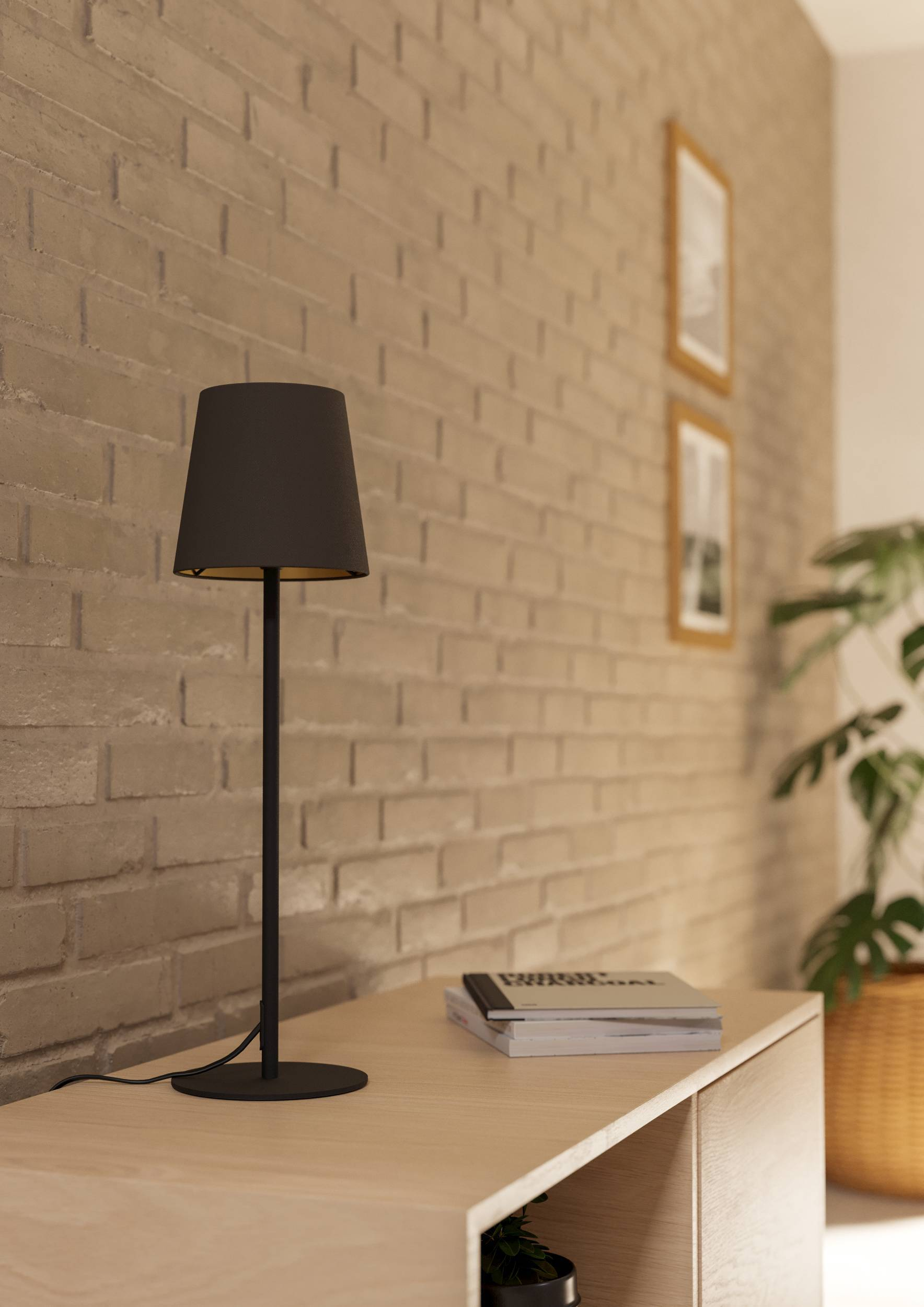 Eine schwarze Lampe mit Lampenschirm steht auf einem Holztisch. Eine Backsteinmauer dient als Hintergrund. Bücher und eine Pflanze sind ebenfalls vorhanden.