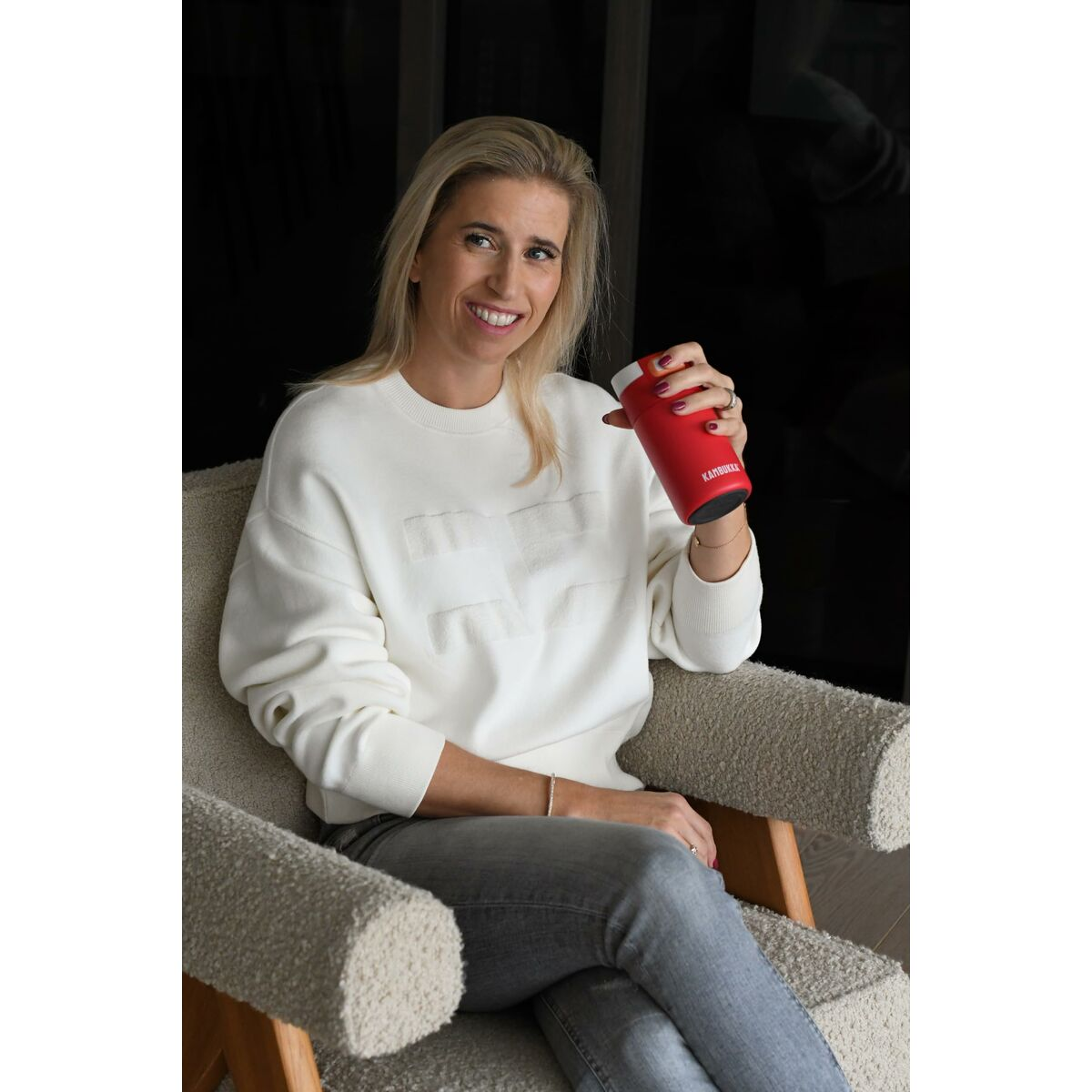 Blonde Frau in weißem Pullover und grauer Jeans, hält einen roten Kaffeebecher, sitzt in einem Stuhl und lächelt.
