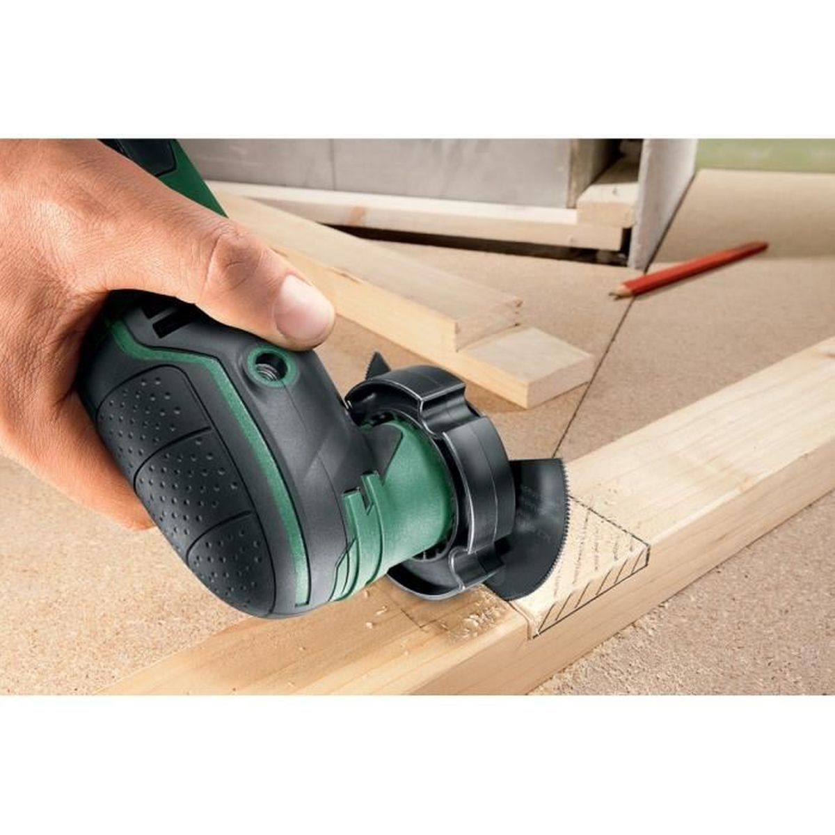 Una mano tiene un utensile elettrico verde e nero che taglia una tavola di legno. Una matita rossa e altri pezzi di legno sono sullo sfondo.