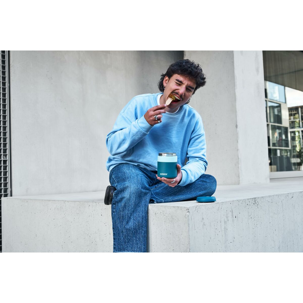 Ein Mann in Blau isst mit einem Holzlöffel und hält einen teal-farbenen Isolierbehälter. Er sitzt auf Beton vor einer grauen Wand.