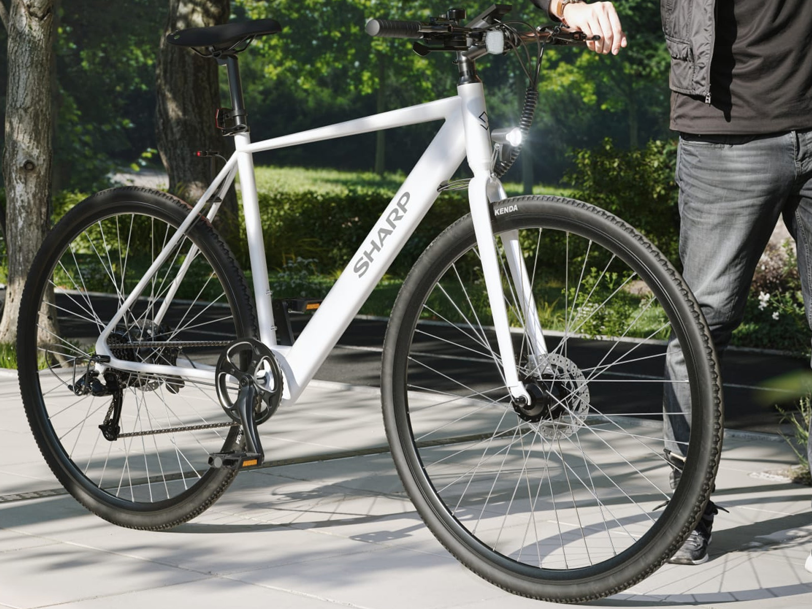 Ein weißes SHARP-Fahrrad ist zu sehen. Eine Person in Jeans und einer schwarzen Jacke hält den Lenker. Bäume sind im Hintergrund.
