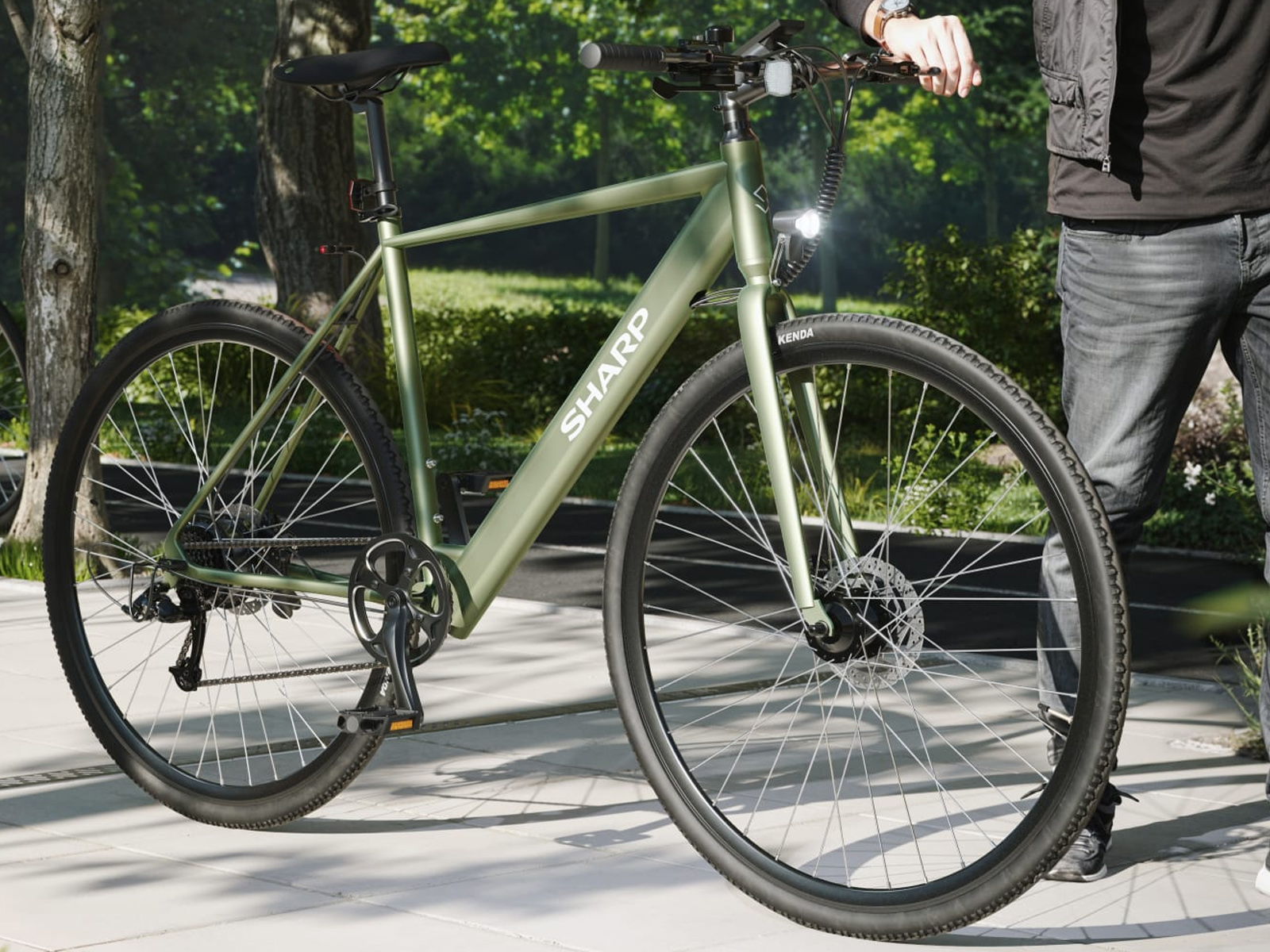 Ein olivgrünes Sharp-Elektrofahrrad wird gezeigt. Eine Person in Jeans hält den Lenker. Bäume im Hintergrund.