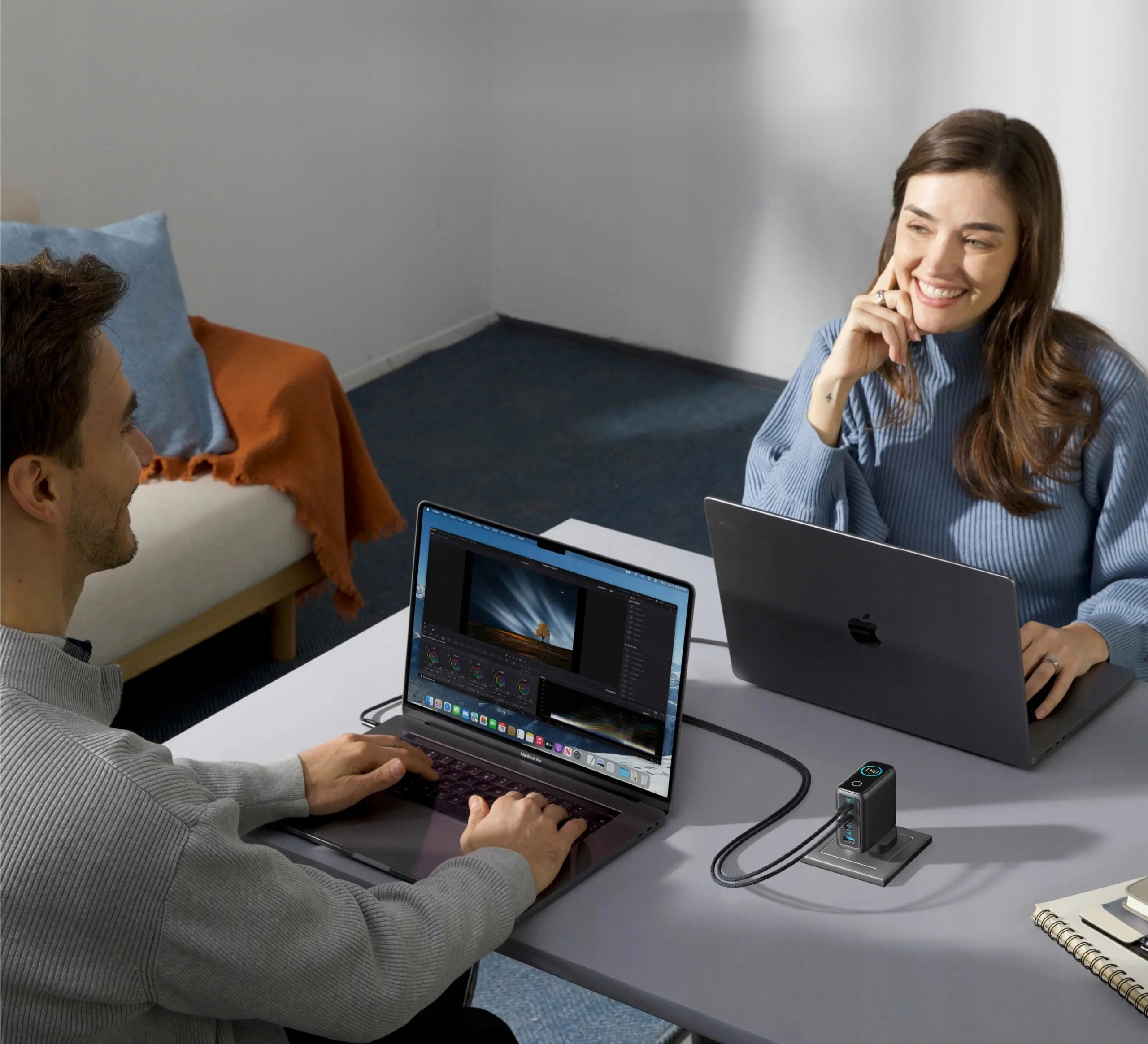 Due persone con laptop su un tavolo. Un caricabatterie è tra loro. Un divano è sullo sfondo.