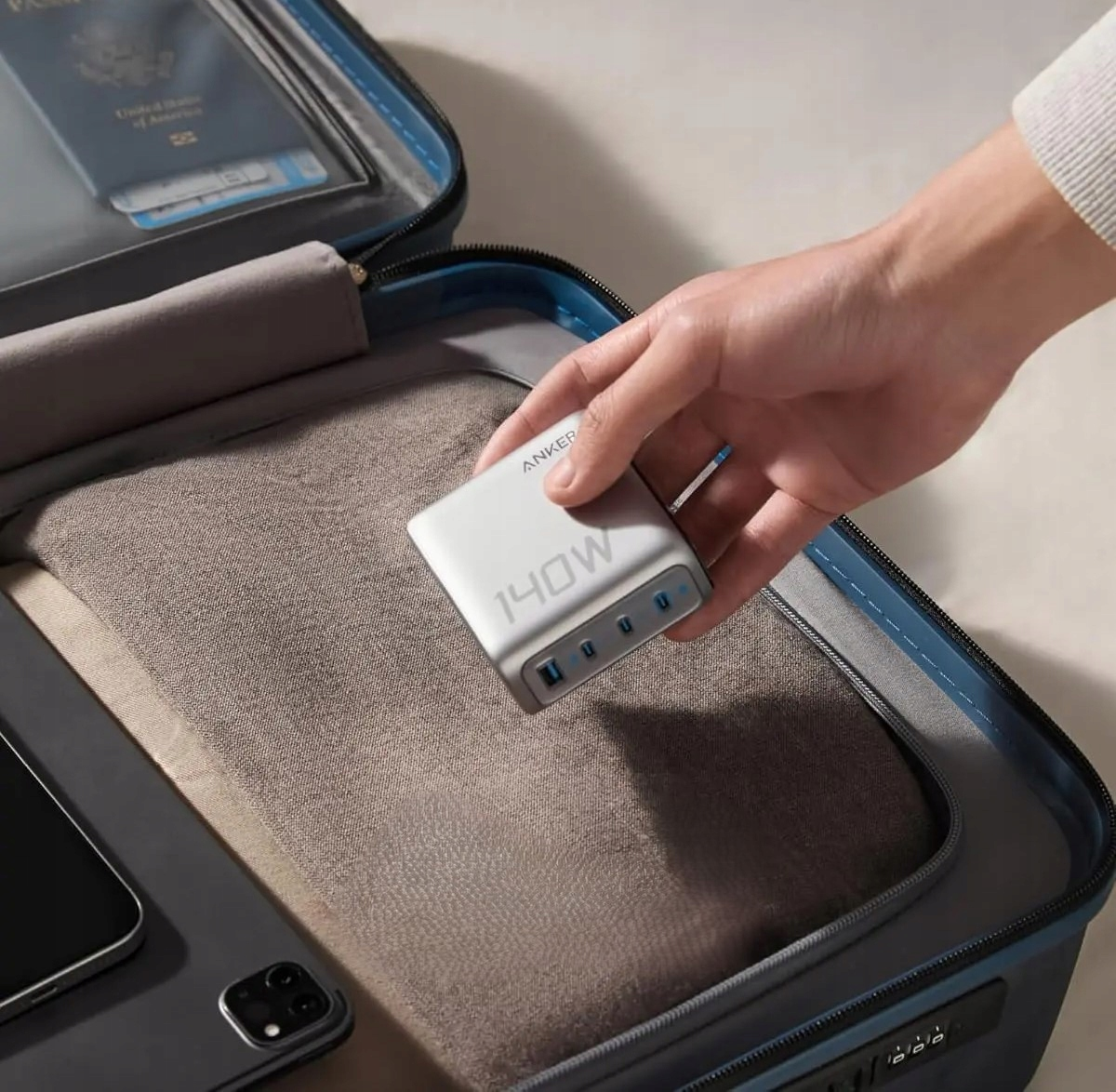 Mano che tiene un caricatore Anker 140W bianco. È in una valigia con un passaporto e un tablet.