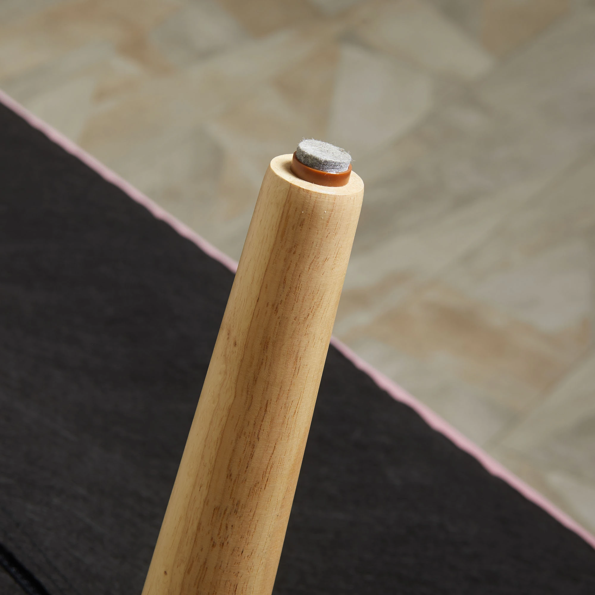 Möbelbein aus Holz mit Filzgleiter. Das Bein ist hellbraun, das Polster grau mit braunem Rand. Schwarze Oberfläche im Hintergrund.