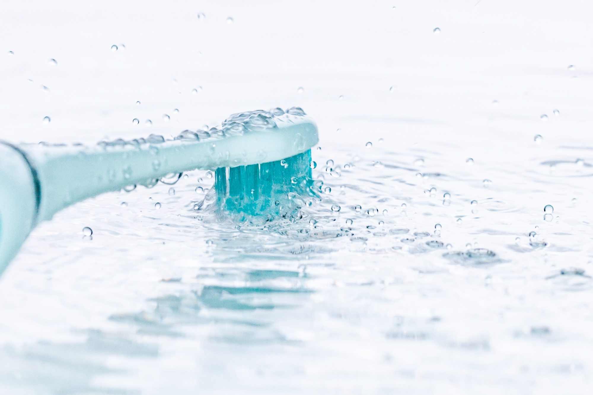 Zbliżenie jasnoniebieskiej główki szczoteczki do zębów zanurzonej w wodzie. Włosie jest turkusowe, z kroplami wody.