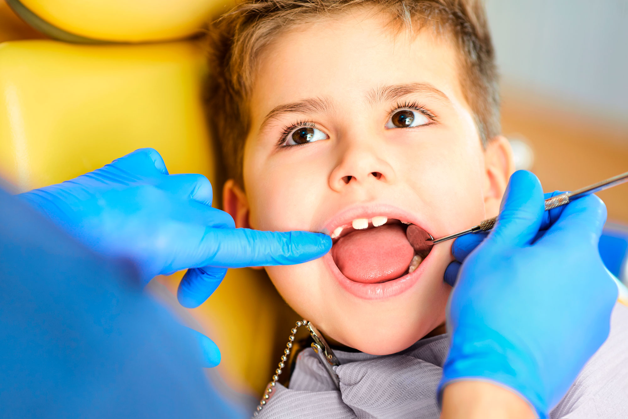 Mały chłopiec u dentysty. Dentysta sprawdza zęby chłopca za pomocą narzędzia, nosząc niebieskie rękawiczki. Chłopiec patrzy w górę.