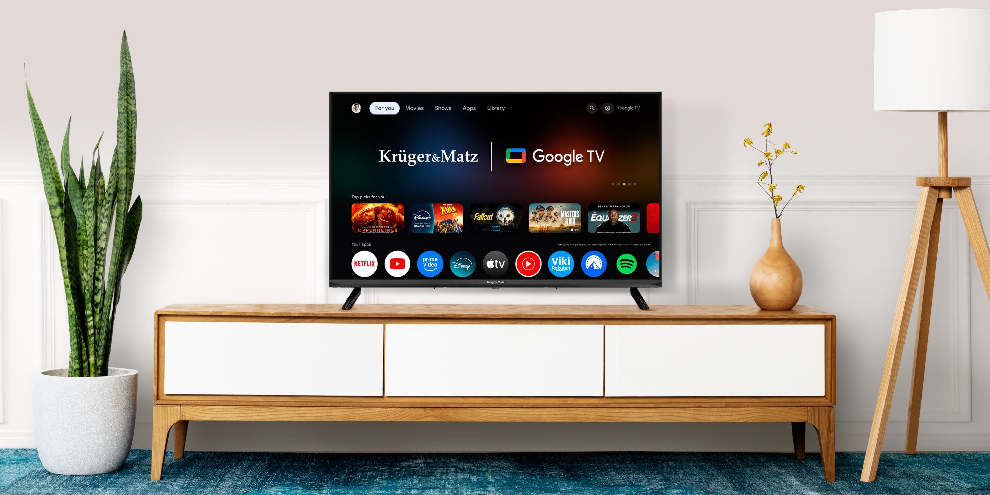 Telewizor wyświetla interfejs Google TV. Stoi na drewnianej szafce. Obok wysoka roślina, lampa i wazon.