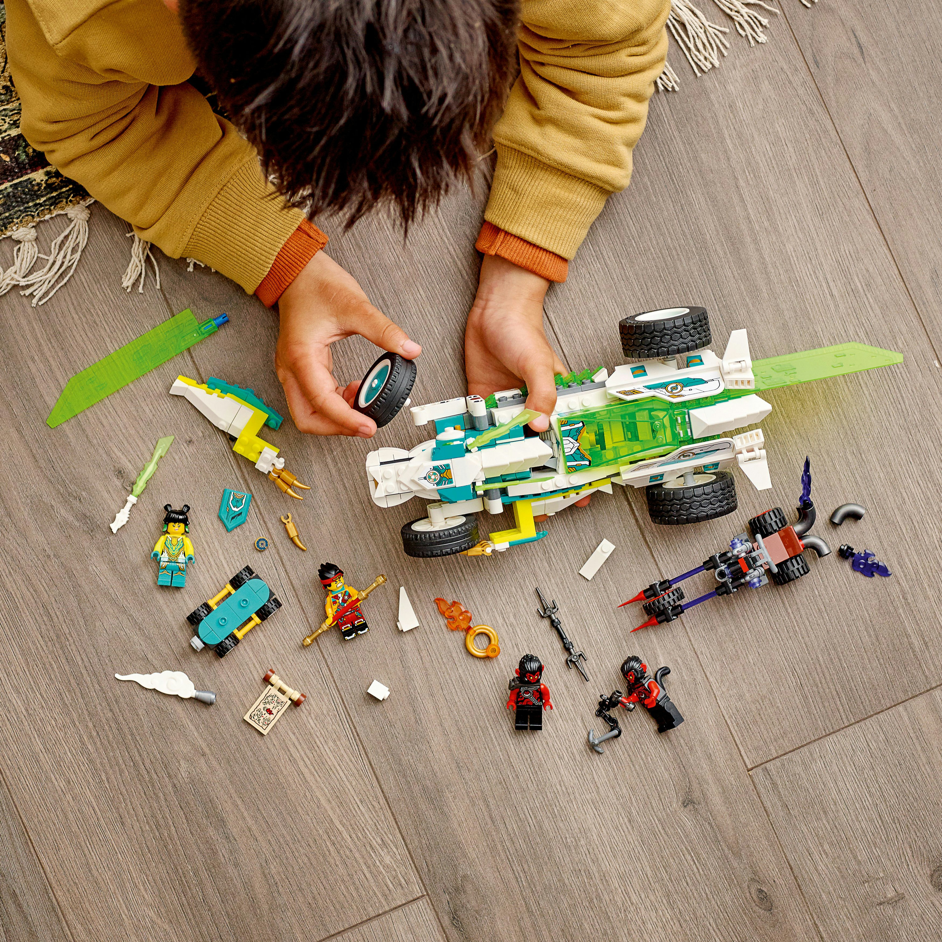 Un bambino assembla un set Lego su un pavimento di legno. Il set include un veicolo, minifigure e vari accessori.