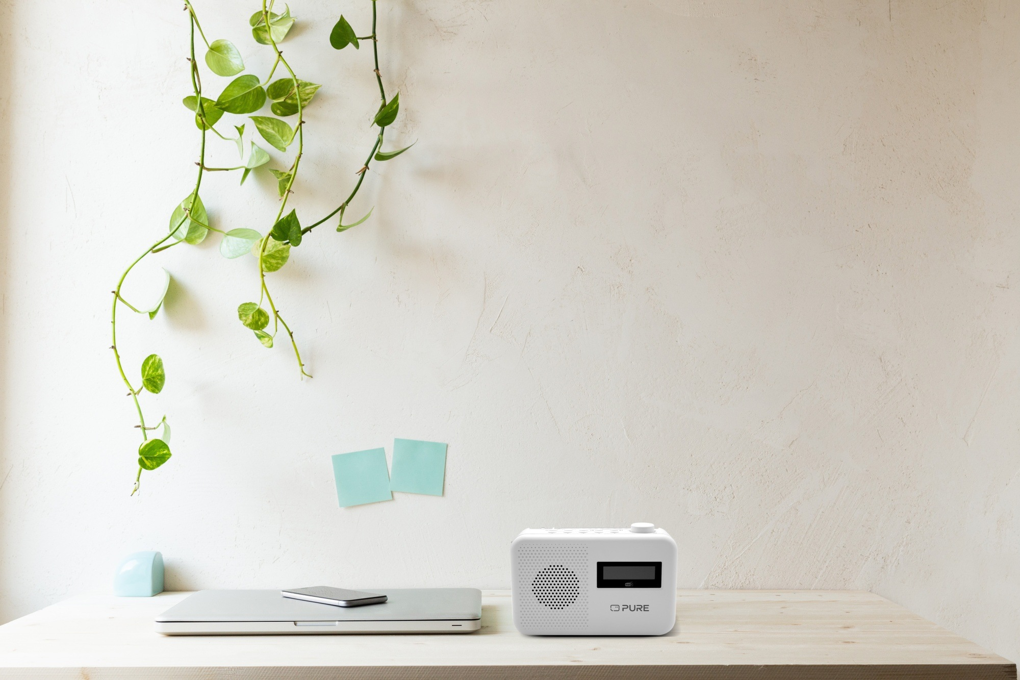 Białe biurko z laptopem, telefonem i radiem. Zielone rośliny zwisają ze ściany. Dwa samoprzylepne karteczki są przymocowane do ściany.