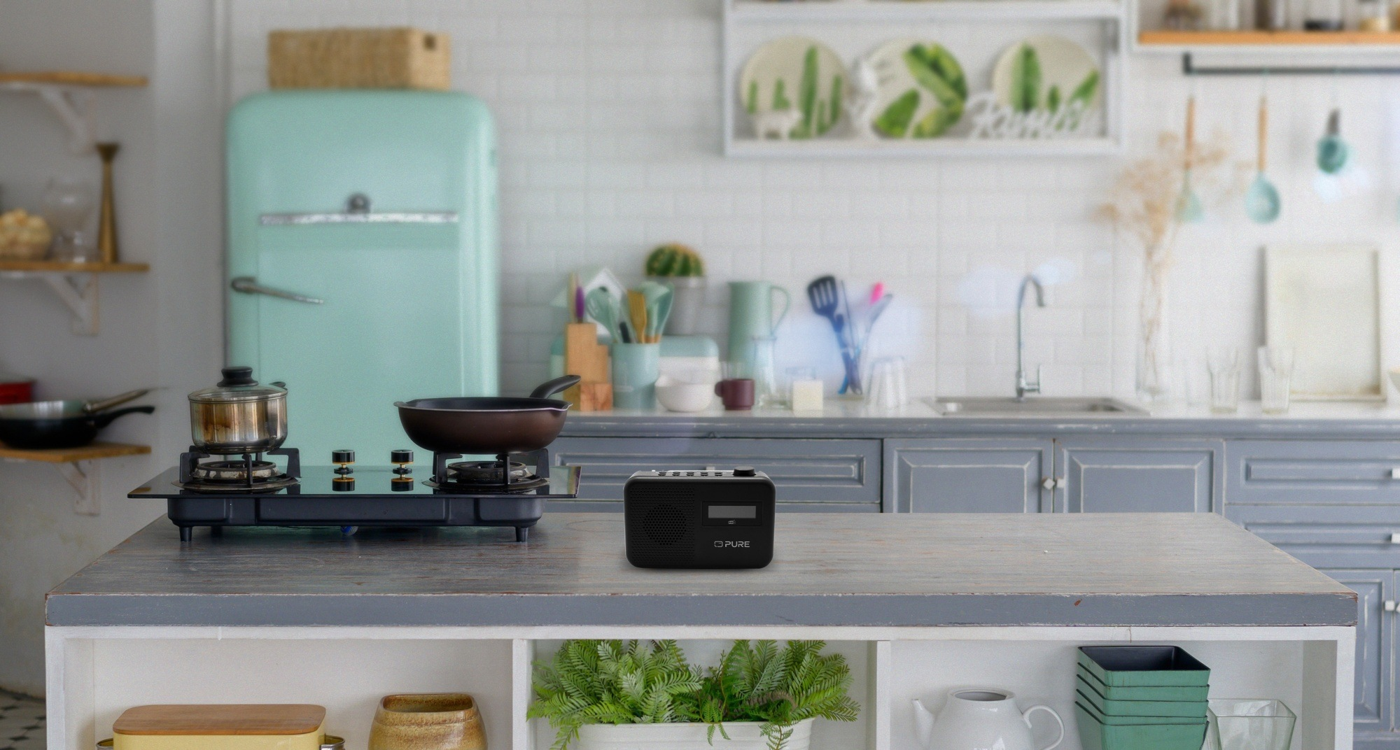 Scena kuchenna z kuchenką, patelnią i radiem. W tle lodówka i półki. Widoczne rośliny i przybory kuchenne.