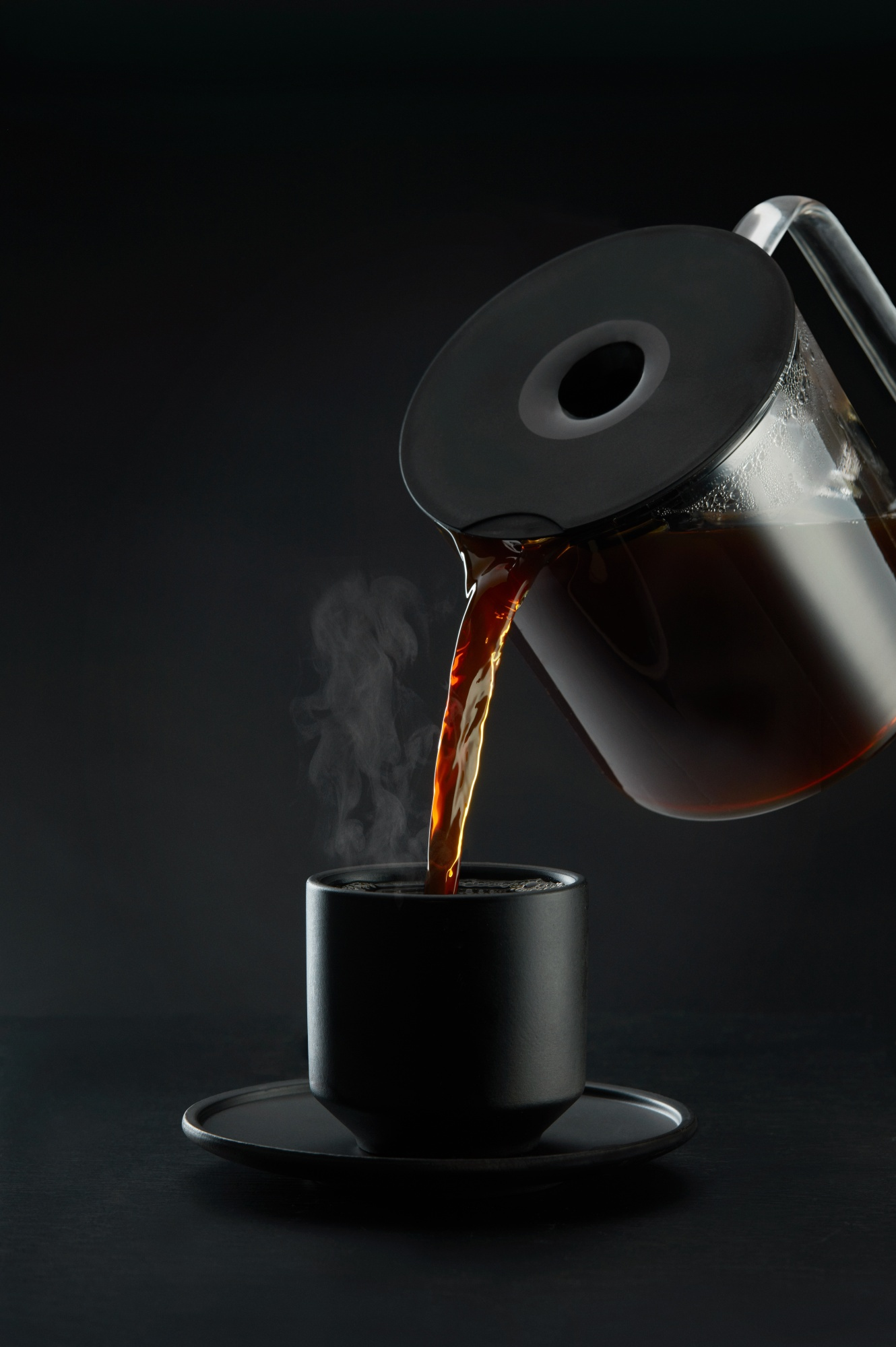 Caffè versato da una caraffa di vetro in una tazza nera. Il vapore sale. La tazza è su un piattino, il tutto su sfondo scuro.