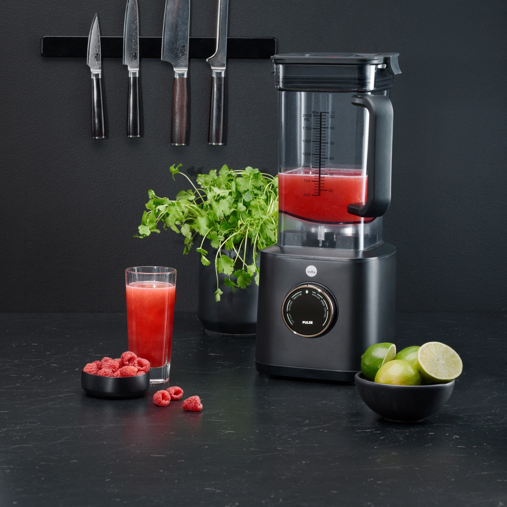 Ein Mixer mit roter Flüssigkeit steht neben einem Glas Saft, Himbeeren, Limetten und Messern. Grüne Kräuter sind ebenfalls vorhanden.
