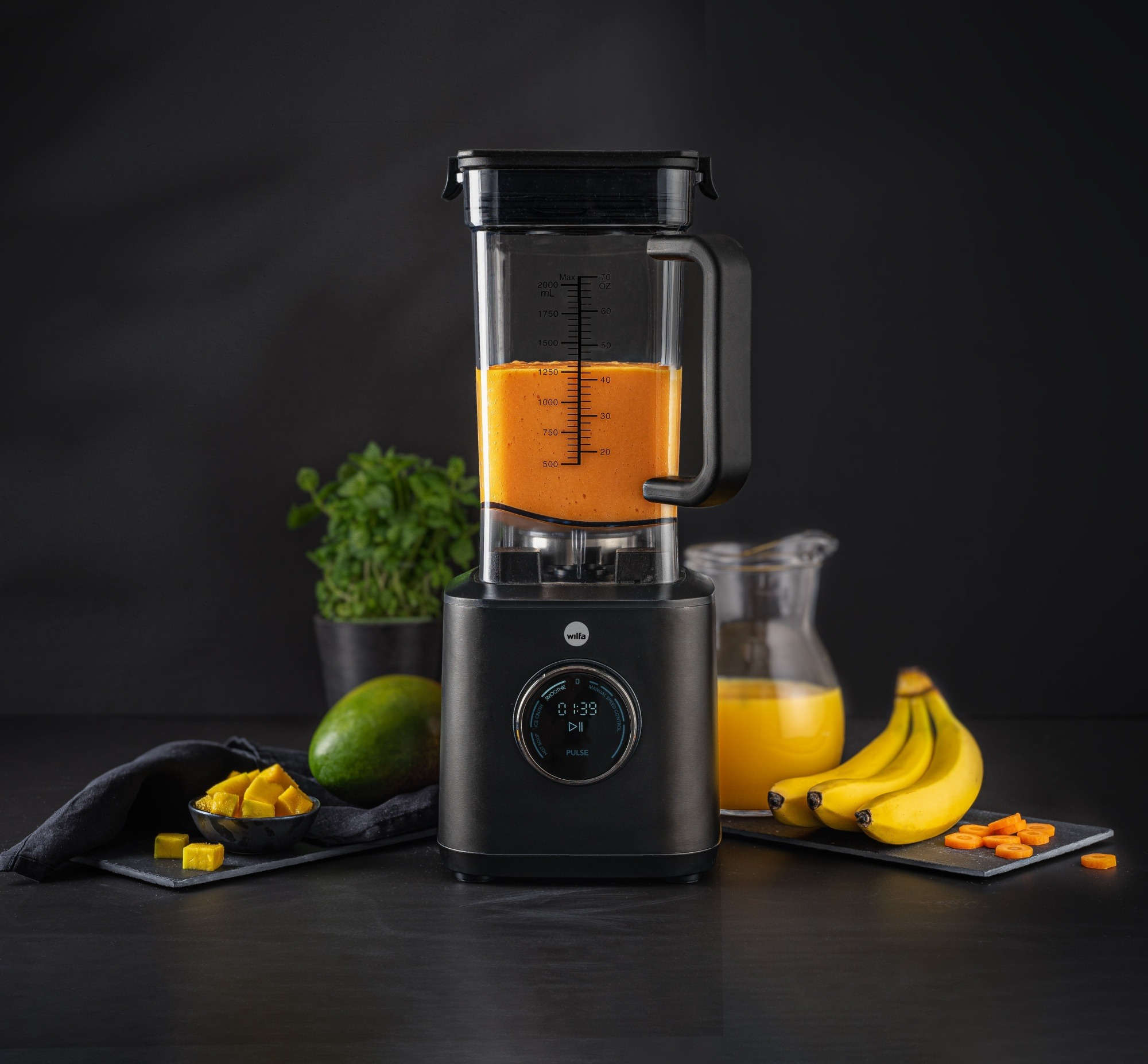 Ein schwarzer Mixer mit orangefarbener Flüssigkeit, umgeben von Früchten wie Mango, Bananen und Karottenscheiben.