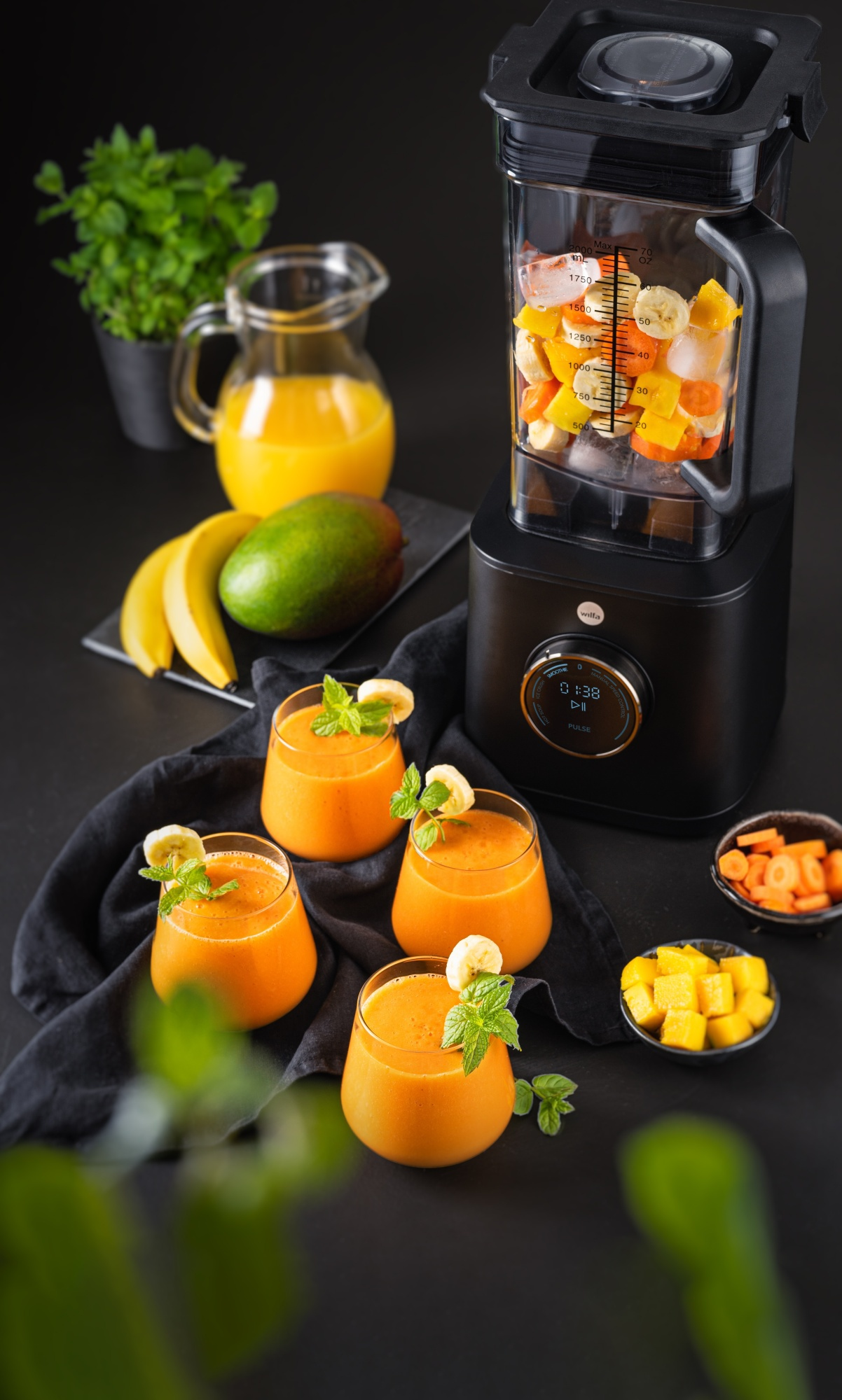 Ein Mixer mit Früchten darin und Gläser mit Smoothie mit Minze und Bananenscheiben. Weitere Früchte und Zutaten sind in der Nähe.
