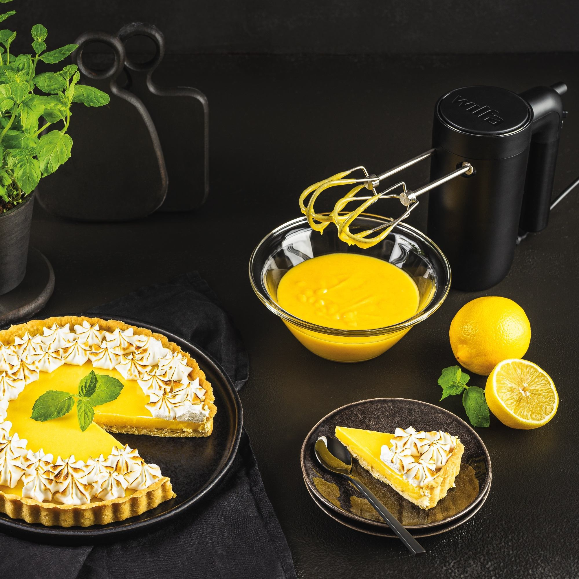 Tarta cytrynowa z bezą z miętą, cytrynami i mikserem na czarnej powierzchni. Kawałek na talerzu.