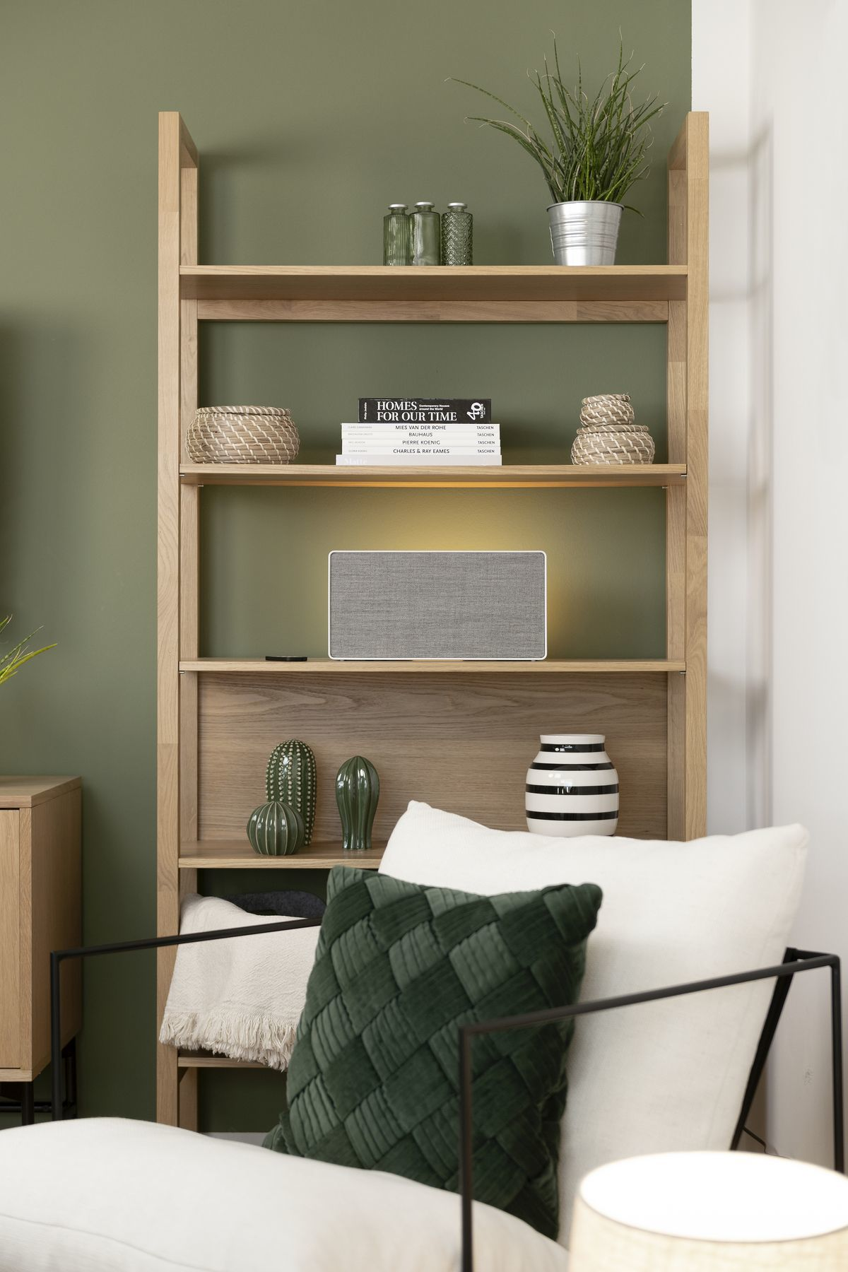 Drewniana półka z roślinami, książkami i dekoracjami na tle zielonej ściany. Poduszki na krześle z przodu.