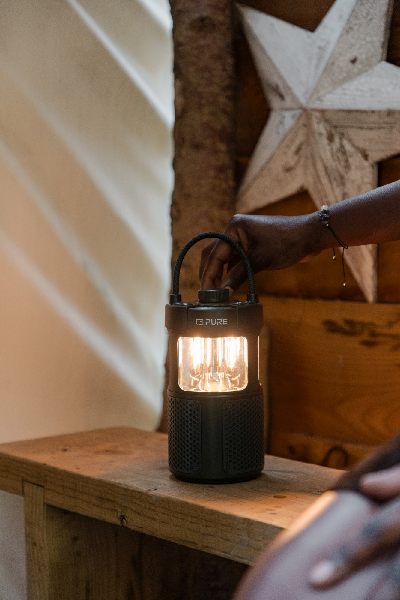 Ręka osoby reguluje zapaloną czarną latarnię na drewnianej ławce. Gwiazda zdobi drewniane tło.