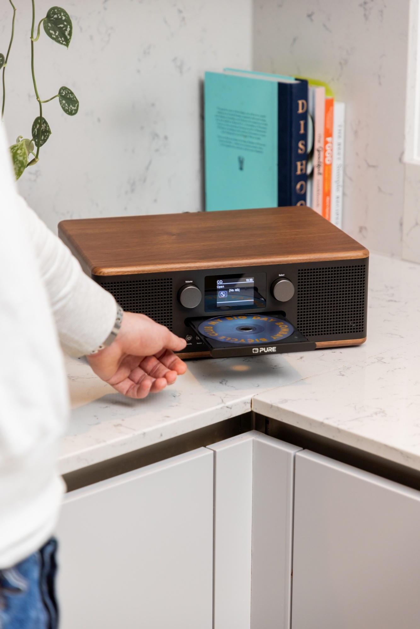 Osoba wkłada płytę CD do drewnianego radia na blacie. Książki i roślina w tle.