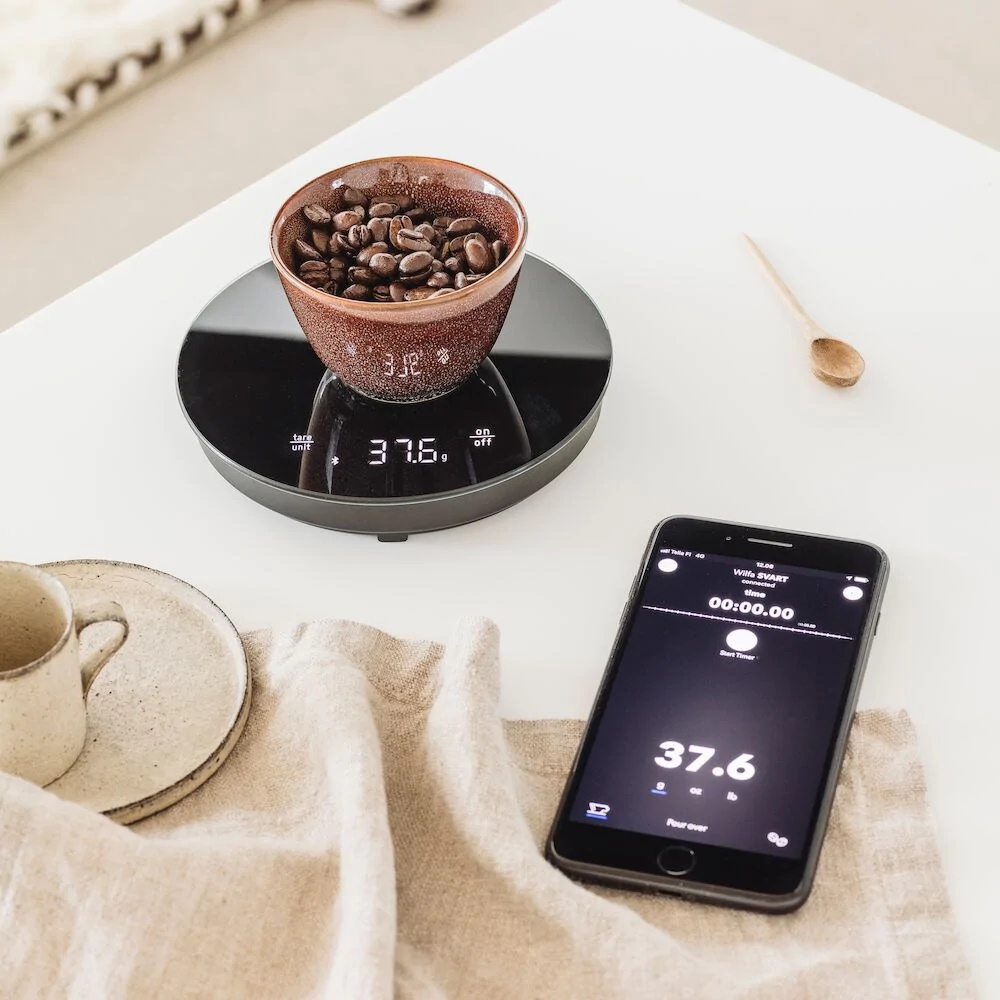 Eine Tasse Kaffeebohnen auf einer Waage mit 37,6. Ein Telefon mit Timer, eine Tasse und eine Untertasse sind ebenfalls vorhanden.