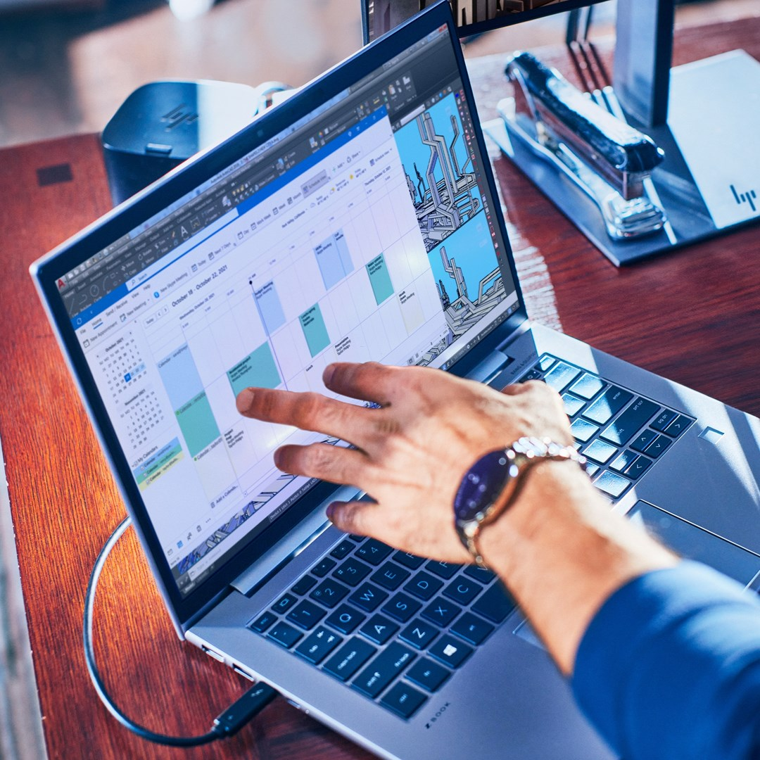 Ręka wskazuje na kalendarz na ekranie laptopa. Laptop stoi na drewnianym stole, w tle zszywacz i monitor.