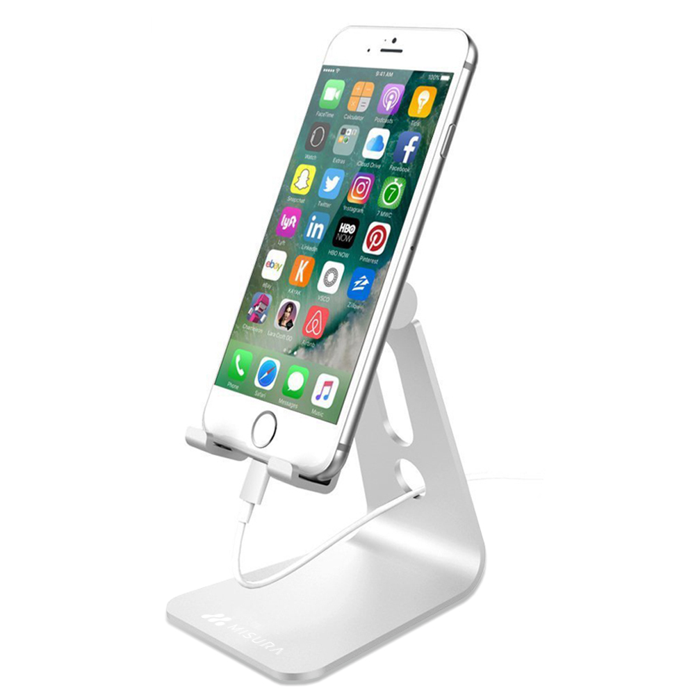 Un iPhone bianco su un supporto argentato. Lo schermo mostra icone di app. Un cavo di ricarica è collegato. Il supporto ha il marchio 'MISURA'.