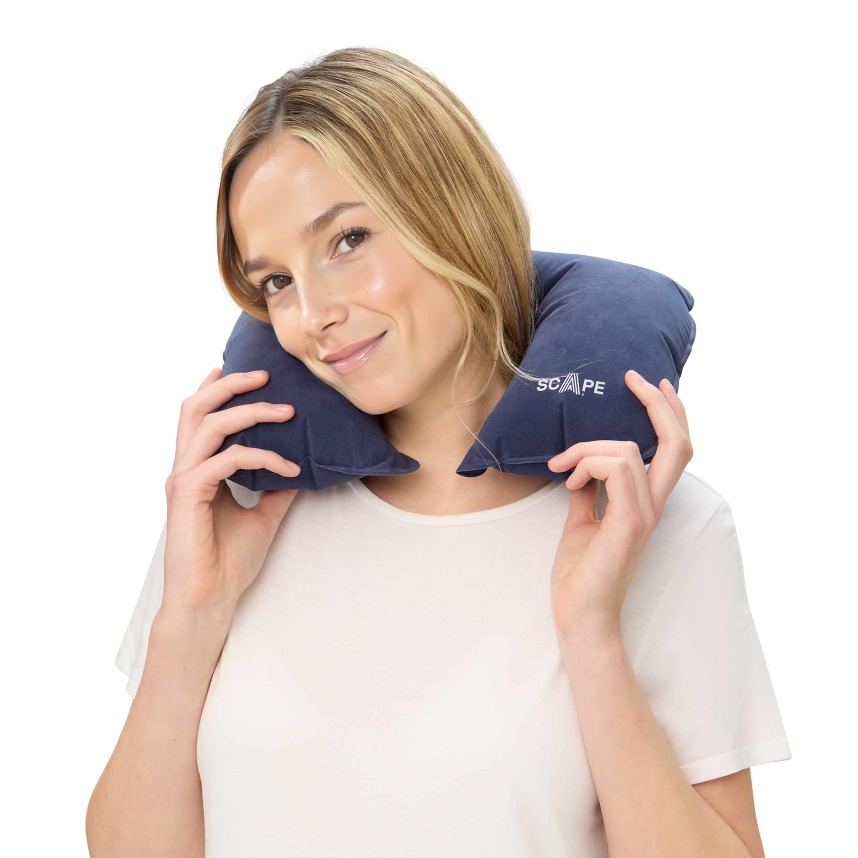 Una mujer rubia sostiene una almohada azul para el cuello. Lleva una camisa blanca y sonríe. La almohada tiene la palabra 'SCAPE'.
